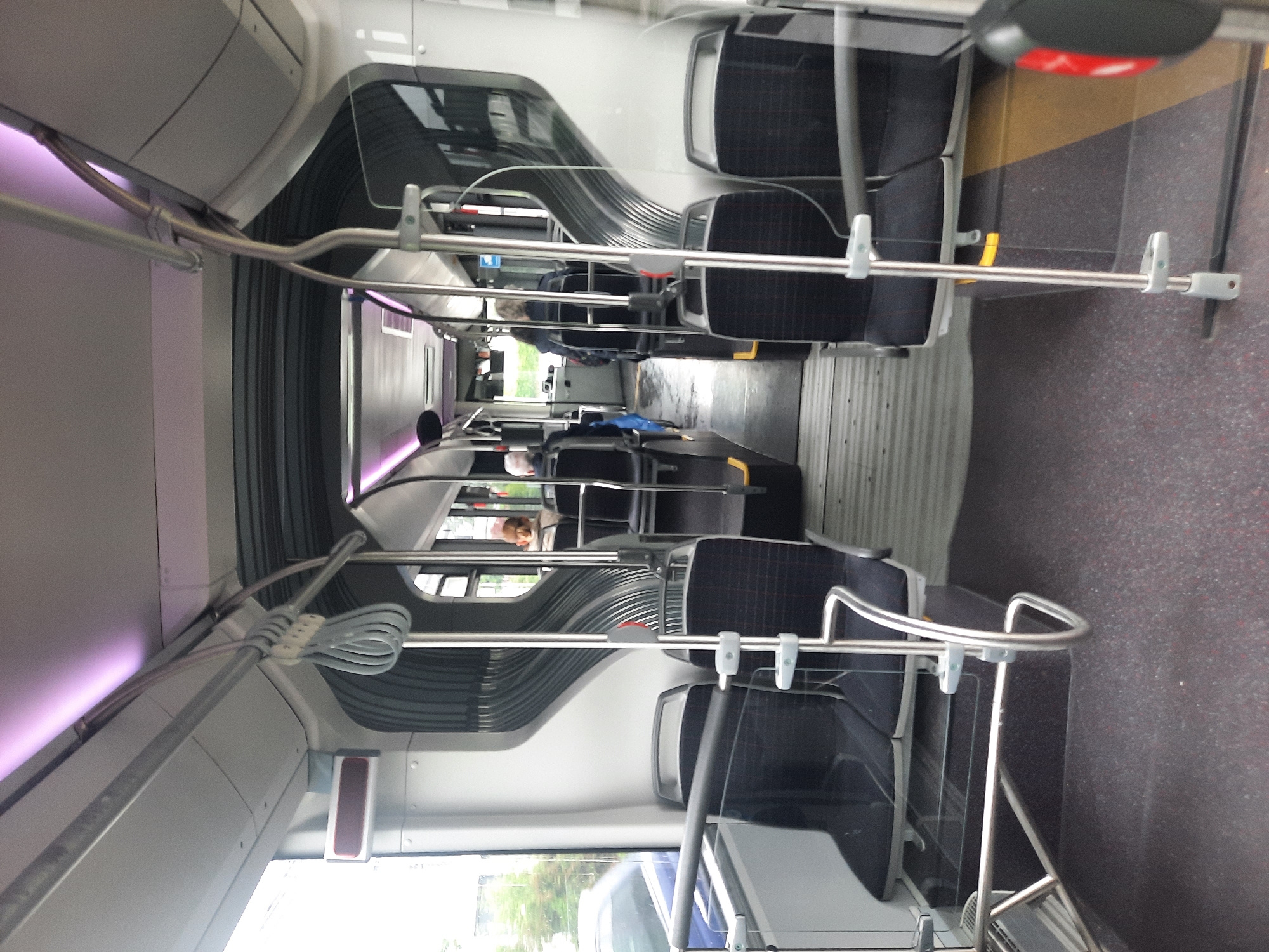 This is an interior view of a modern bus, possibly a public transport vehicle, looking towards the rear. The seats are black and arranged in a 2x2 configuration on both sides of a central aisle. The aisle flooring is dark gray with red speckles. Overhead, there are metal handrails and a lighting system that emits a purple hue. Various people are visible sitting in the seats further down the aisle, with their backs to the camera. One individual in the foreground on the left is wearing a blue top. The bus appears to be stationary, with windows showing an exterior view of trees and a car. The design of the bus suggests it is relatively new, with sleek, modern lines and materials. Without further context, it is not possible to definitively locate this bus in Rosenheim, Germany, as the image only provides an interior view of a vehicle that could be present in many cities.