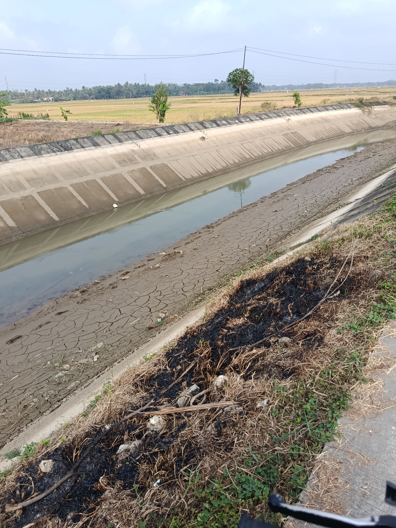 The image captures a scene along a concrete canal in Bantarsari, Indonesia. The canal, with its textured concrete walls, carries a moderate amount of murky water. The banks of the canal are a mix of dry, cracked earth and patches of dry grass, some of which appear to have been burned, leaving behind dark, charred debris. Beyond the canal, a flat landscape unfolds, characterized by cultivated fields, likely rice paddies, with a line of trees and scattered buildings in the distance. The sky is partly cloudy, suggesting daytime. There are no people or animals immediately visible in the frame.