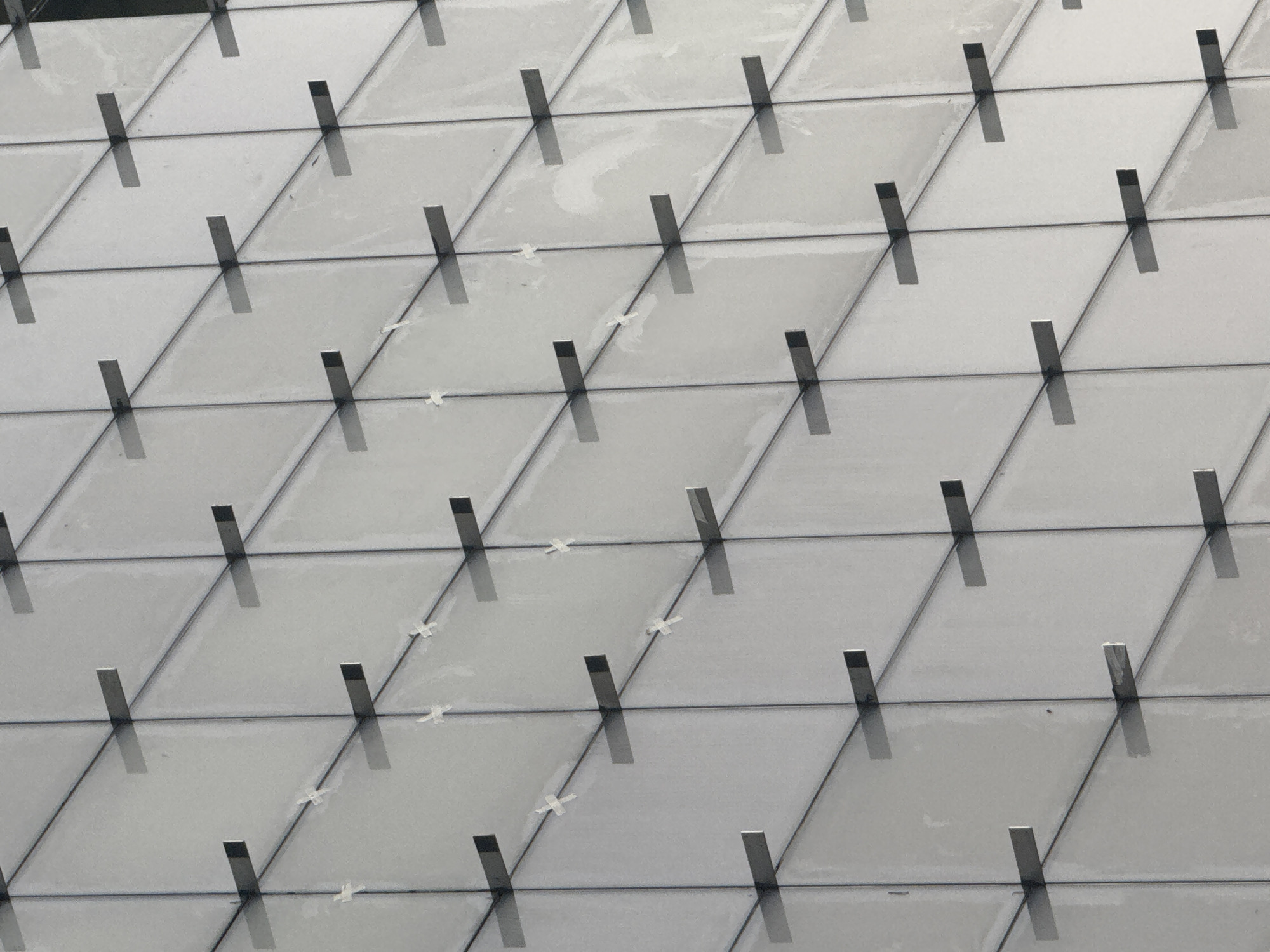 The image shows a close-up view of a building facade, likely in Tel-Aviv, Israel, based on architectural styles found there. The facade is constructed from triangular or diamond-shaped panels, seemingly made of a light-colored, possibly opaque, material. Each panel is intersected by a dark, slender, vertical element, likely a metal brace or support. These vertical elements create a regular grid pattern across the facade. Some of the panels show signs of minor staining or discoloration. Small pieces of white adhesive or residue are visible at the corners of some of the panels. The overall impression is of a modern, geometric design.