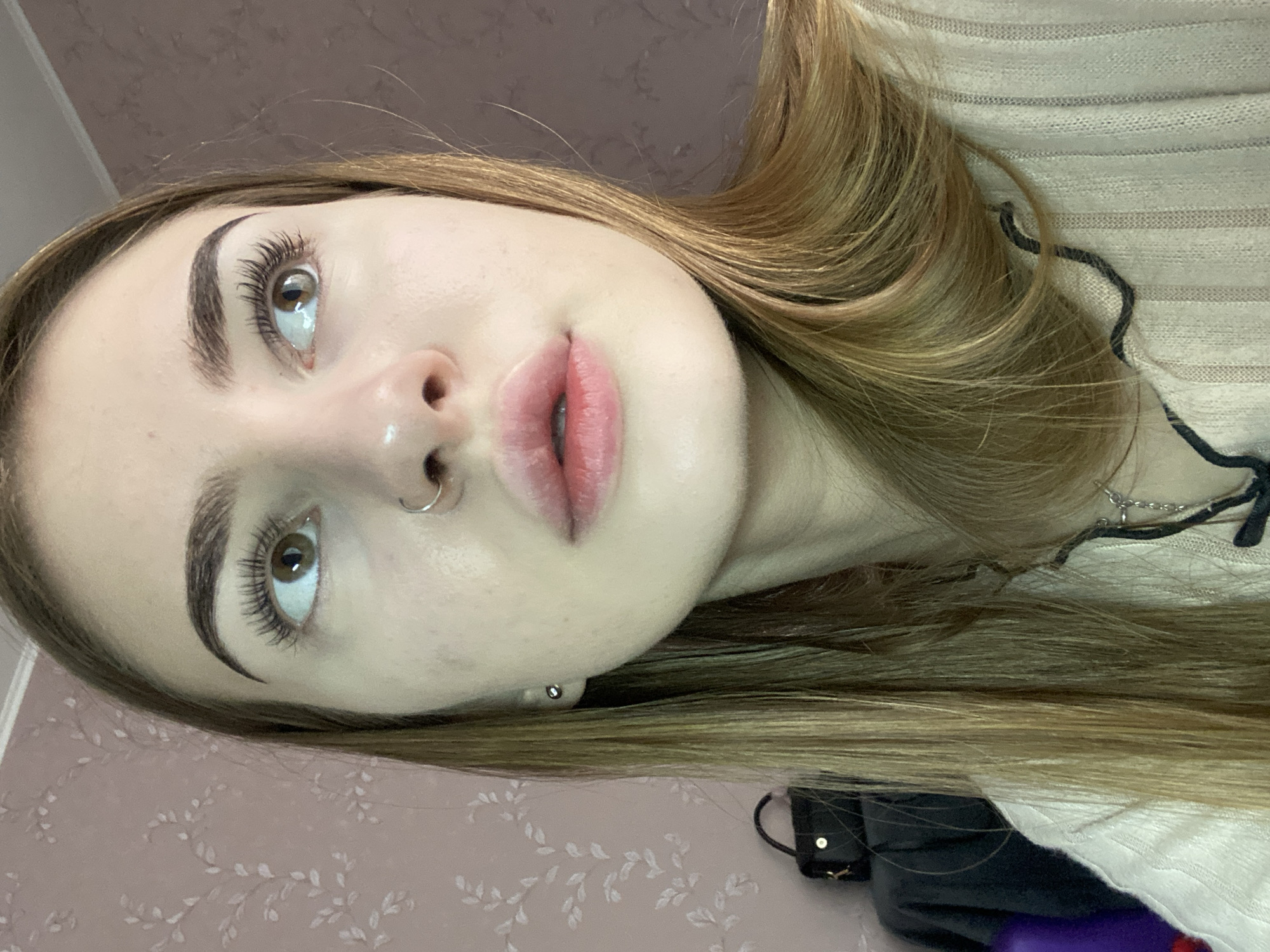 The image shows a close-up, slightly upward-angled shot of a young woman's face and upper torso, taken indoors. She has light skin, full lips with a pink tint, and brown eyes looking towards the upper right. Her eyebrows are dark and well-defined, and her eyelashes are long and dark. She has a small silver hoop nose ring and a small silver stud earring in her visible ear. Her blonde hair is long and straight, falling over her shoulders. She is wearing a light-colored, ribbed knit sweater. Behind her, the background consists of a pink patterned wallpaper and a dark draped object, possibly a piece of furniture or clothing, with a black purse on it. The overall lighting is soft and appears to be from an indoor source. There is no indication or visual information to suggest the location is in the city of Дьяково, Russia.