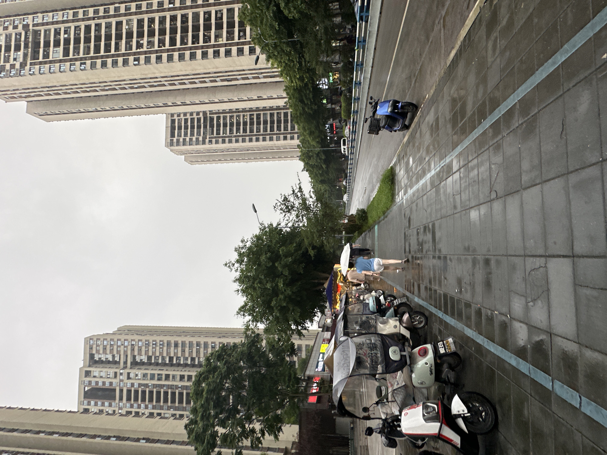 The image depicts a street scene in Chongqing High-tech Zone, China, under overcast skies and light rain. A sidewalk paved with dark grey tiles, wet from the rain, runs along the right side of the frame. A blue electric scooter is parked on the sidewalk. On the left, a row of electric scooters and three-wheeled vehicles are parked. Several people are walking on the sidewalk, some carrying umbrellas. In the background, tall apartment buildings line the street, interspersed with trees. The street itself is wet and shows some traffic.