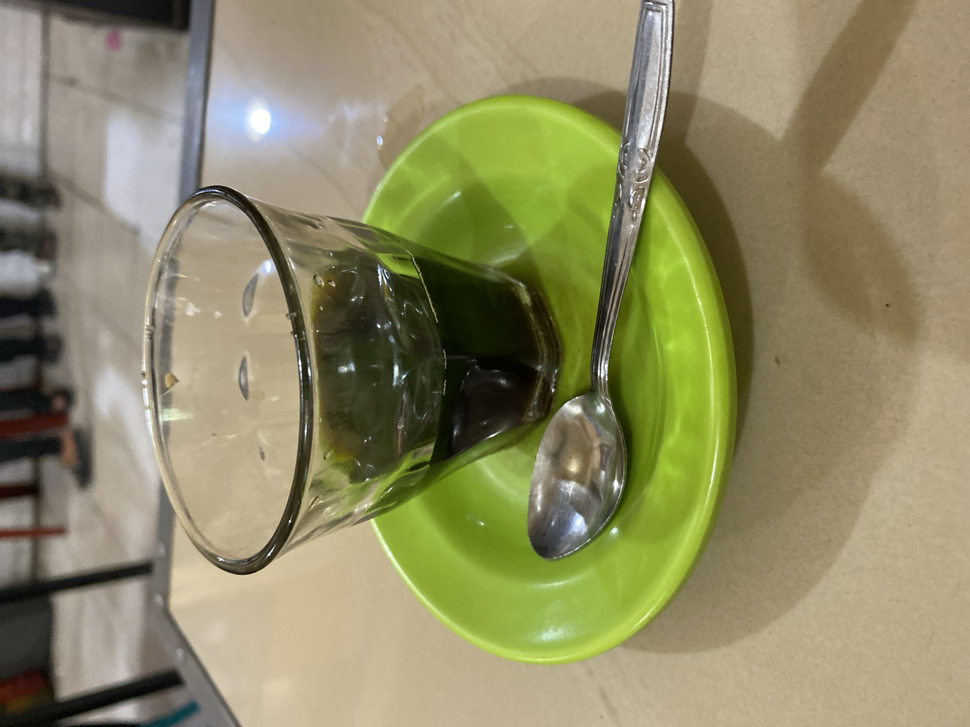 A close-up, slightly elevated shot captures a glass of dark liquid, possibly coffee or tea, resting on a bright green saucer. A silver spoon lies horizontally across the saucer, its handle pointing towards the right edge of the frame. The glass has a slight ripple texture, and the liquid fills the bottom third of the glass. Reflections of the environment are visible on the glass and the spoon. The saucer is a vibrant, solid green, with a subtle texture. The background is out of focus, showing a light-colored table surface and hints of people's legs and footwear, suggesting an indoor setting, potentially a cafe or restaurant in Lhokseumawe, Indonesia, as indicated by context. The lighting is moderate, casting a soft shadow from the glass and saucer onto the table.