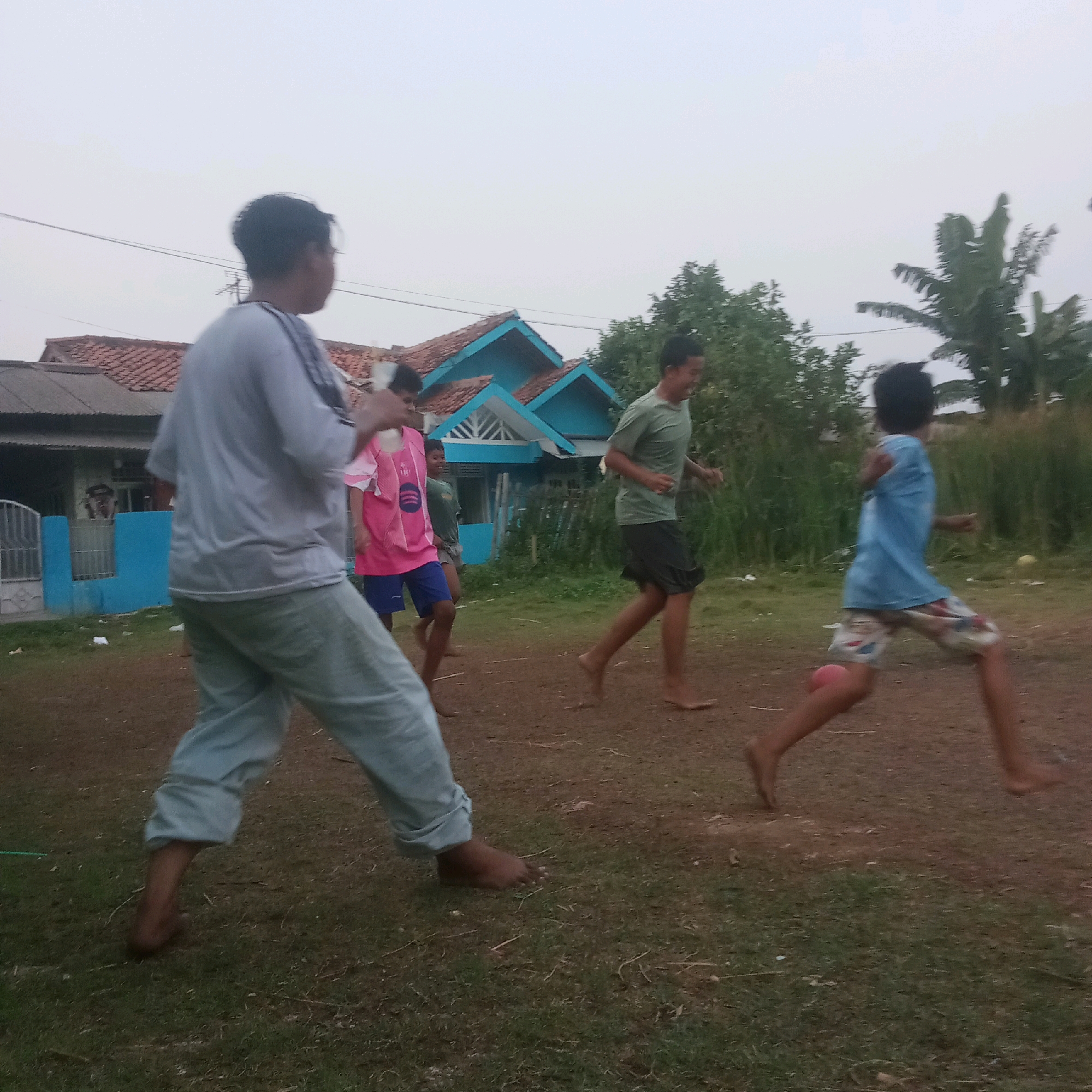 The image depicts an outdoor scene, likely in Karawang, Indonesia, where four individuals are engaged in a game of Sepak Takraw. The sky is overcast, suggesting it could be late afternoon or early morning. The ground is a mix of dirt and grass.
In the foreground on the left, a person wearing a light gray t-shirt and light blue pants is captured mid-stride, with their left foot raised and bent, demonstrating an active pose. Their right foot is planted on the ground.
To their right, another person is wearing a bright pink t-shirt with a circular logo and dark blue shorts. This individual is also in motion, with their body turned to the left, and their right leg extended.
Further to the right and slightly behind, a person in a green t-shirt and dark shorts is running, with their left leg bent and their right leg extended. Their head is turned to the left, and they appear to be smiling.
On the far right, a person in a blue t-shirt and patterned shorts is running, with their body angled to the left. Their left leg is extended, and their right leg is bent. They are barefoot.
In the background, a residential structure with a reddish-brown tiled roof and blue accents on the roofline is visible. Trees and other vegetation surround the area, with some appearing to be palm trees, consistent with the Indonesian landscape. A metal fence and a blue structure are also present near the house. The overall atmosphere suggests a casual recreational activity taking place in a semi-urban or rural residential setting.