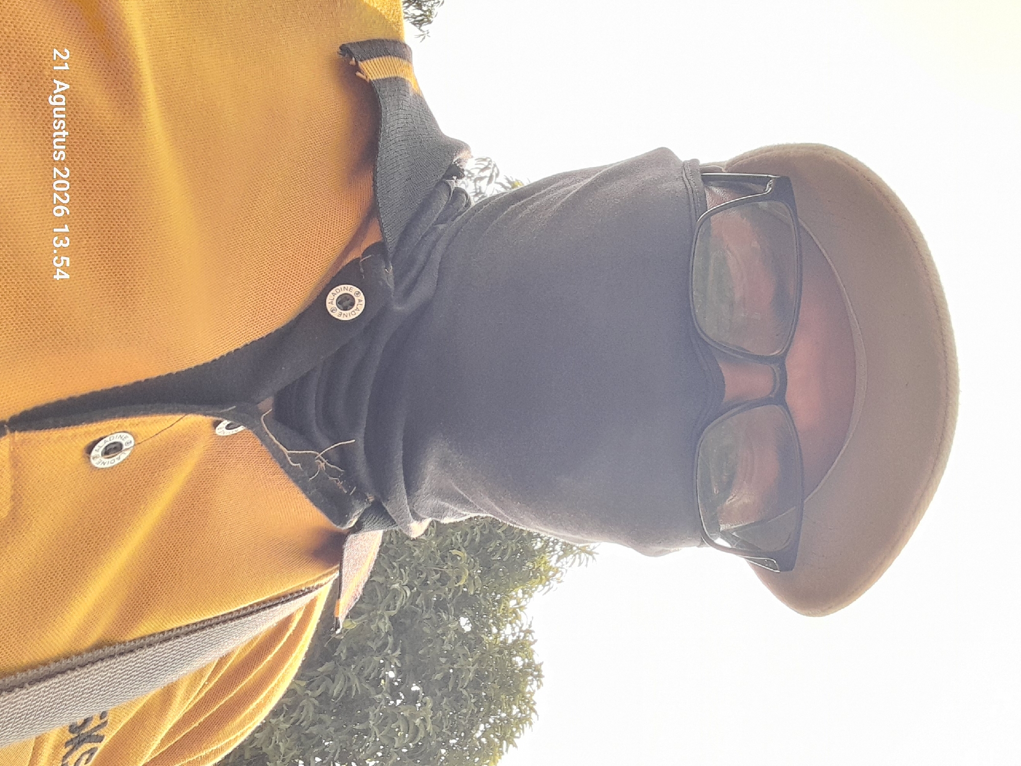 A low-angle, close-up selfie depicts a person from the chest up, wearing a tan beret, dark-rimmed glasses, and a black face covering. The person is also wearing a yellow polo shirt with a dark collar and trim, adorned with two circular buttons, one of which displays the text "ALADINE" and "MINE". A grey strap is visible across the right shoulder. The background consists of a bright, overcast sky and green foliage, suggesting an outdoor setting. The timestamp "21 Agustus 2026 13.54" is overlaid on the bottom left of the image, indicating the photograph was taken on August 21, 2026, at 1:54 PM. The location is specified as Takeran, Indonesia.