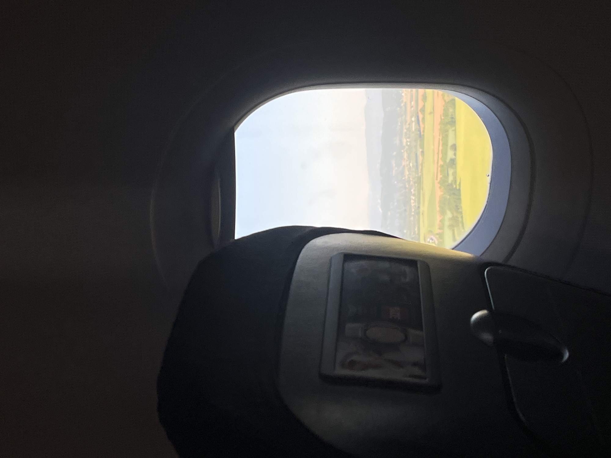 The image depicts a view from an airplane window. Outside, the landscape shows a mixture of green fields, vegetation, and possibly some urban or residential areas in the distance. The sky appears bright and clear. Inside the aircraft, the back of a seat with a screen is visible. Based on the landscape and sky, it is possible that the location could be near Colombier-Saugnieu, France, though this is not definitively proven by the image alone.