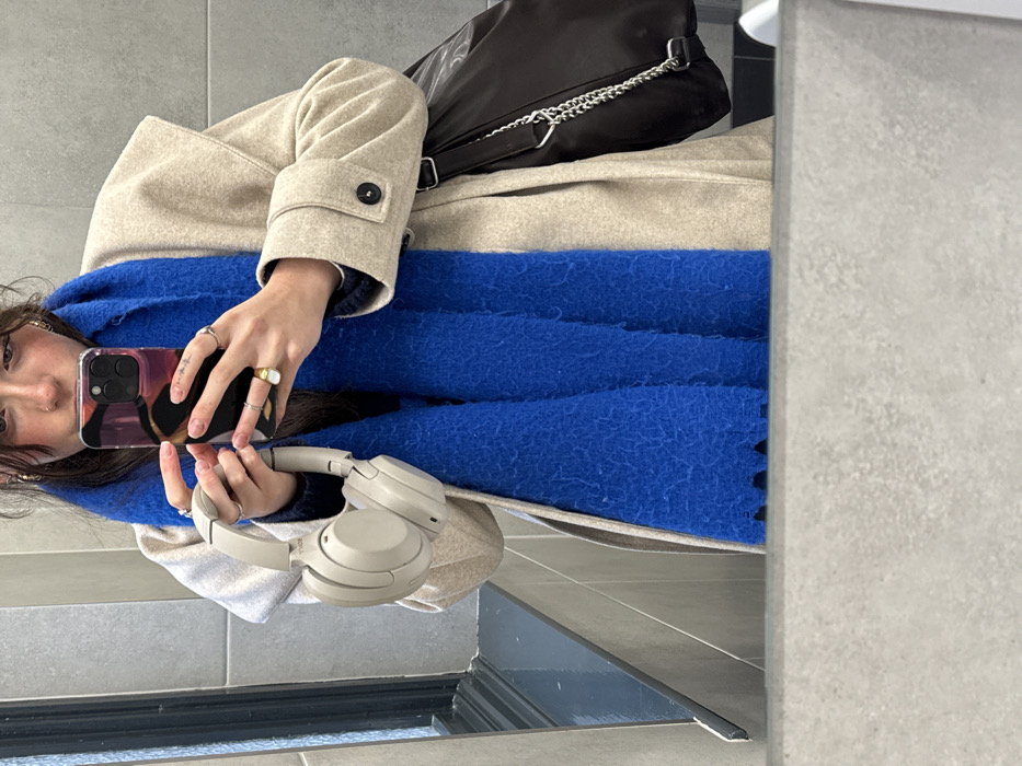 A mirror selfie reveals a person wearing a beige coat and a large, fluffy blue scarf. A black handbag with a silver chain strap rests on the coat. The person holds a phone in one hand and a pair of beige headphones in the other. Multiple rings adorn their fingers. The selfie captures the person's face partially, showing dark hair and some facial features. The background includes grey tiling.