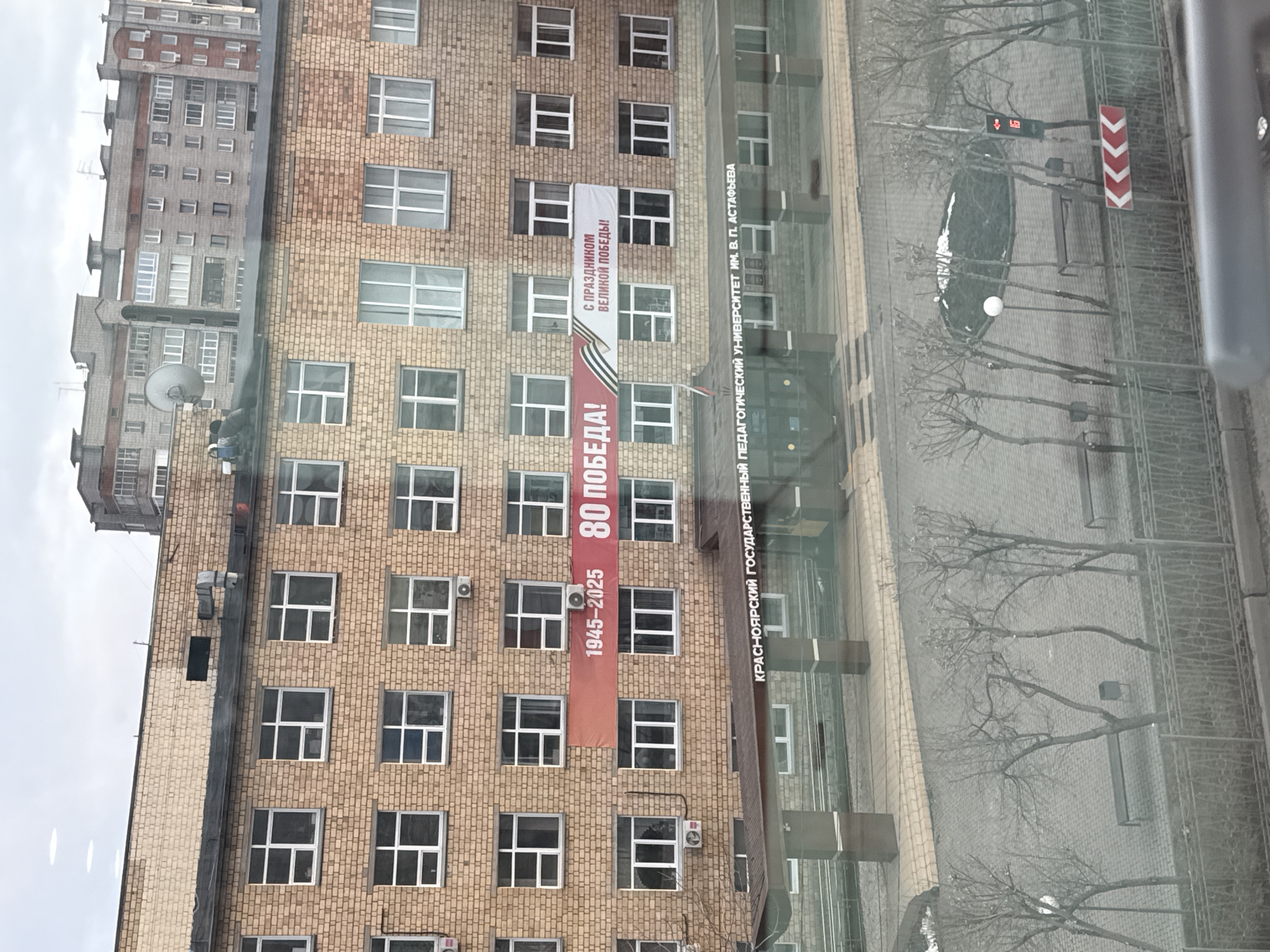 A medium, eye-level, indoor shot of a view outside of a window depicts a brick building with a banner hanging on the side, trees, and the street in front of the building in the city of Krasnoyarsk, Russia. The building is tall, likely over ten stories high, and made of brown bricks, with many white-framed windows. There is a vertical banner hanging between a few windows on the right side of the building that is read "С ПРАЗДНИКОМ ВЕЛИКОЙ ПОБЕДЫ!" and “1945-2025 80 ПОБЕДА!”. Next to the building, a sign reads "КРАСНОЯРСКИЙ ГОСУДАРСТВЕННЫЙ ПЕДАГОГИЧЕСКИЙ УНИВЕРСИТЕТ ИМ. В. П. АСТАФЬЕВА”, which translates to Krasnoyarsk State Pedagogical University named after V. P. Astafieva. The trees on the street are thin and bare. The street is gray with a traffic light in the distance.