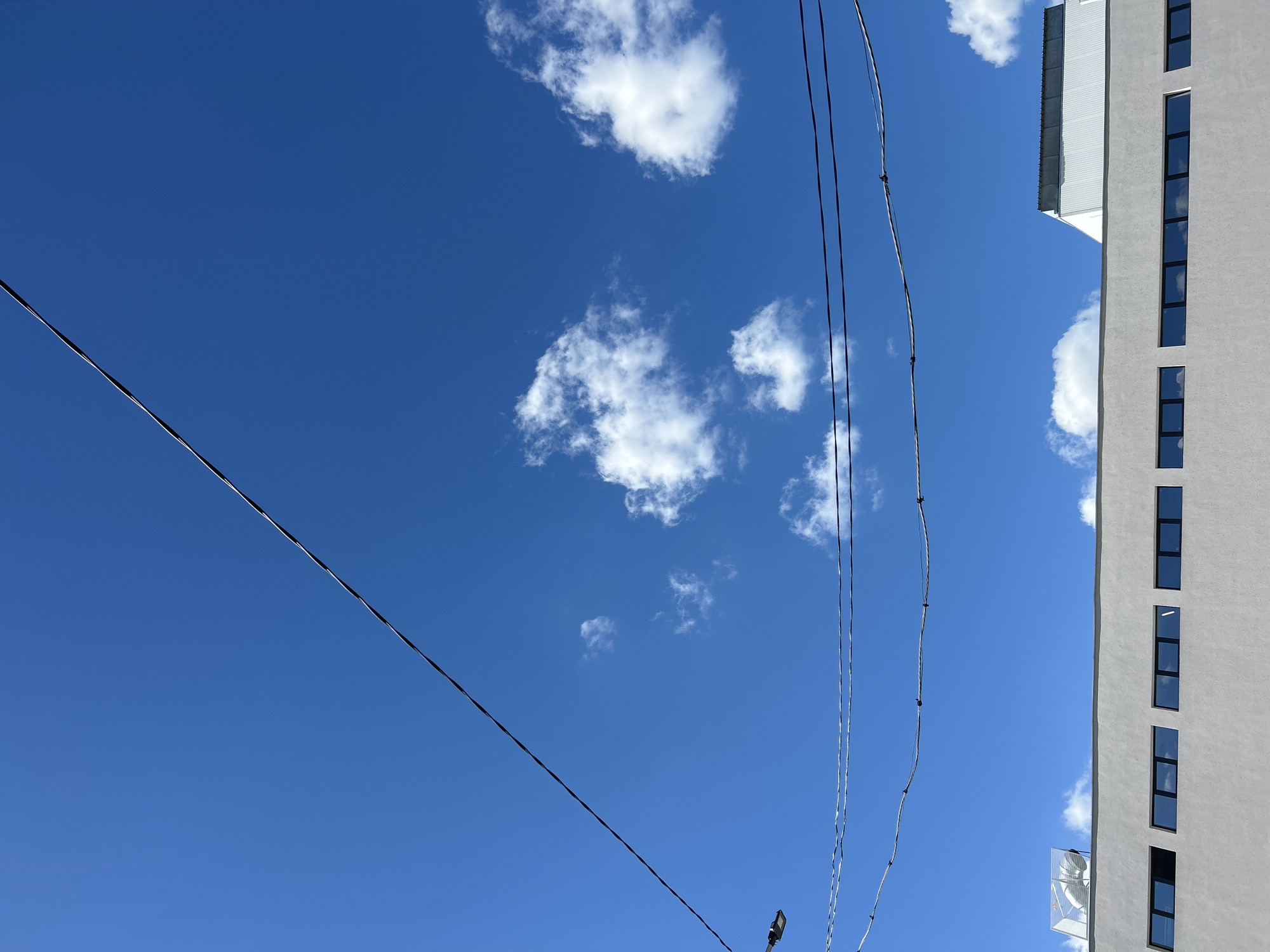 A clear, bright blue sky with scattered white clouds forms the upper portion of the image. Several power lines stretch across the sky, one diagonally from the top left to the middle right, and two others horizontally in the lower middle. A street lamp is visible on the left side. The lower part of the image shows the top of a modern, light gray building with a long row of rectangular windows. The windows reflect the sky and clouds. On the roof of the building, towards the left, is a white ventilation unit enclosed in a metal cage. To the right of that, a portion of a white building with a corrugated metal roof is visible. The scene appears to be an urban or industrial setting on a sunny day.
