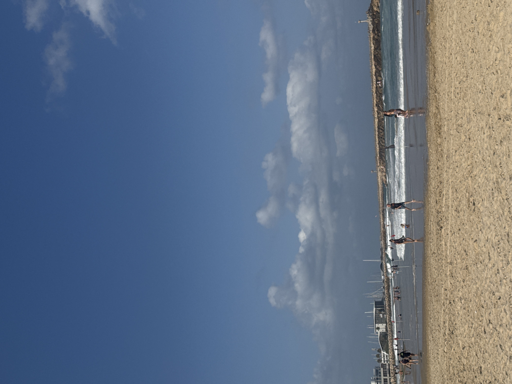 The image depicts a lively scene at Herzliya Beach in Herzliya, Israel. The foreground is dominated by coarse, golden sand, indicative of a typical beach environment. In the middle ground, several individuals are engaged in various activities on the wet sand and in the shallow water. Some are walking along the shoreline, while others appear to be wading or playing in the waves. The water is a vibrant turquoise, with moderate waves breaking.
Beyond the immediate beach area, a long, rocky breakwater stretches across the frame, dotted with lights and a lighthouse at its far end, marking the entrance to a harbor. In the distance, a modern urban landscape is visible, with buildings and numerous sailboats docked in a marina. The sky above is a clear, bright blue, punctuated by scattered white clouds. The overall atmosphere suggests a warm, sunny day with recreational activities in full swing.