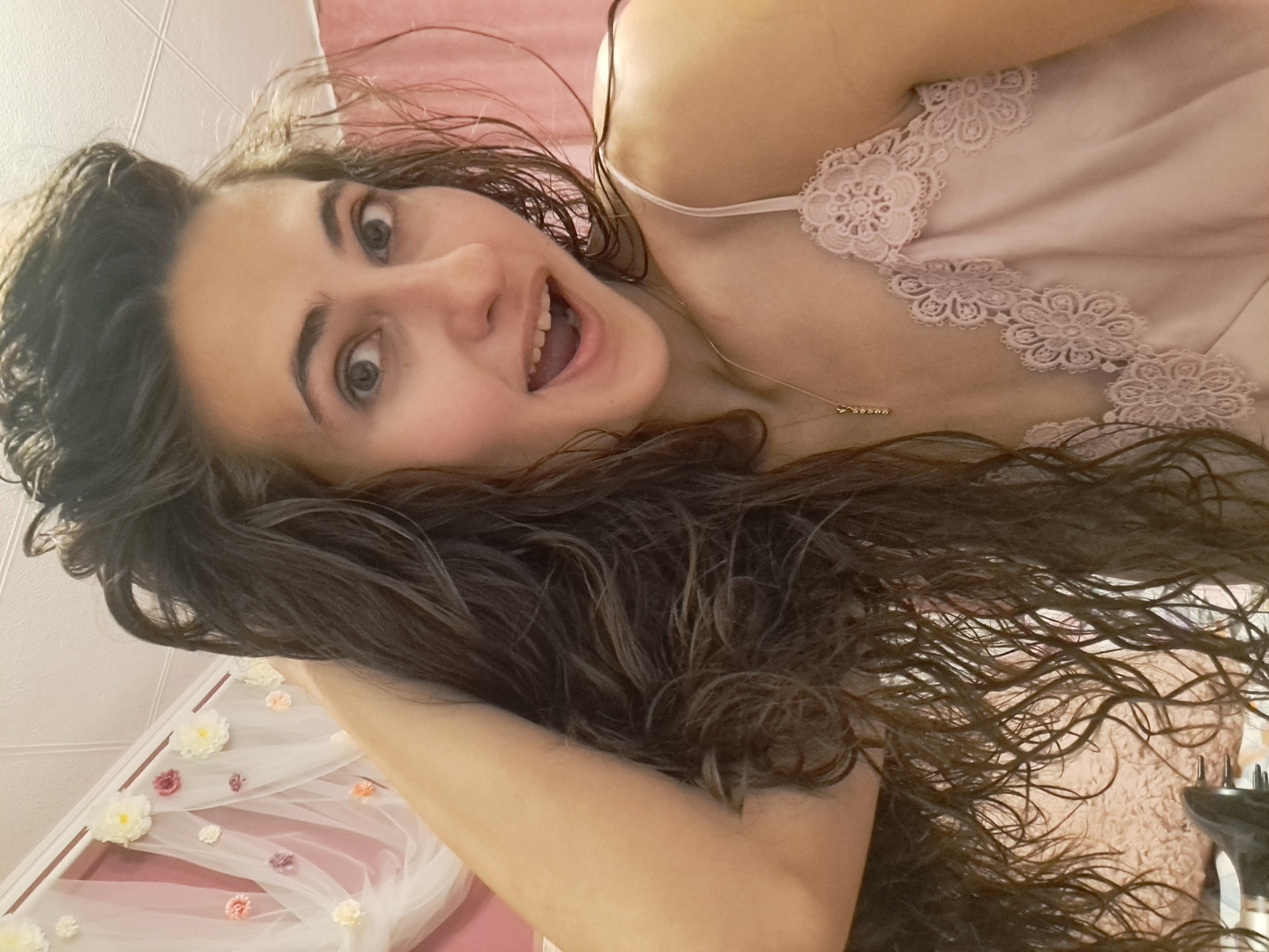 A young woman with wet, dark brown hair is shown from the chest up, looking at the camera with an open mouth. She is wearing a light pink camisole with lace trim. Behind her, a sheer pink fabric with attached white and pink flowers is visible, creating a decorative backdrop. The lighting suggests an indoor setting, possibly a bedroom. There is no indication of the location being in Negovan, Bulgaria.