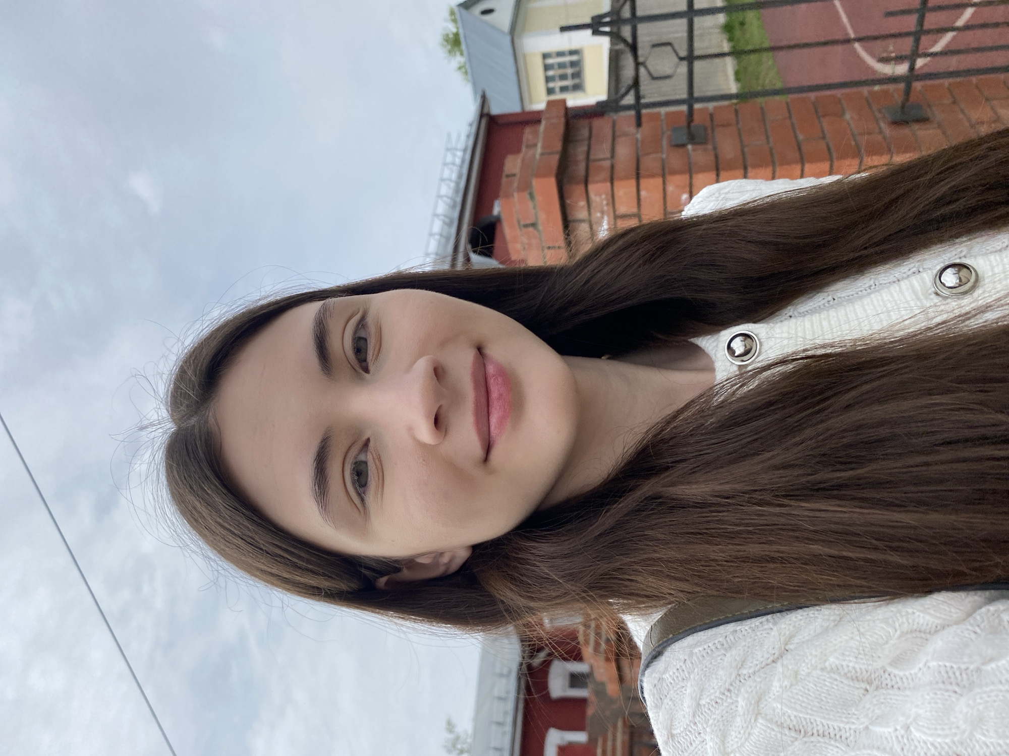 A selfie taken by a young woman with long, brown hair. She is wearing a white, knitted top with silver buttons. Behind her is a red brick building with white window frames, partially obscured by a black iron fence. Beyond the fence, a red surface with white lines is visible, potentially a sports court. The sky is overcast. While it is difficult to pinpoint the exact location in Votkinsk, the architectural style of the brick building suggests it could be near older sections of the city.