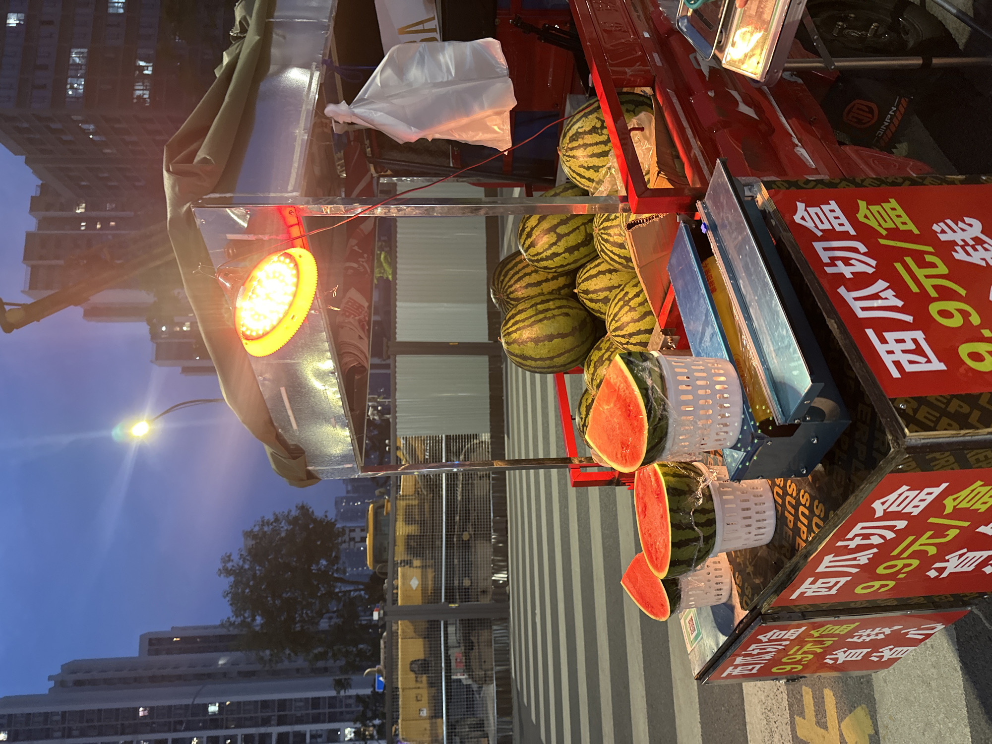 The image depicts a street vendor's cart selling cut watermelons in the Chongqing High-tech Zone, China. The cart is red and is situated on a pedestrian crossing at dusk. Multiple watermelons are visible, both whole and pre-cut and packaged. Red signs with Chinese characters, indicating "watermelon cut box" and a price of "9.9 yuan/box," are displayed prominently. The vendor's cart is illuminated by a bright yellow overhead lamp. In the background, tall apartment buildings and construction equipment are visible, suggesting an urban environment under development. The sky is a deep blue, indicative of twilight.