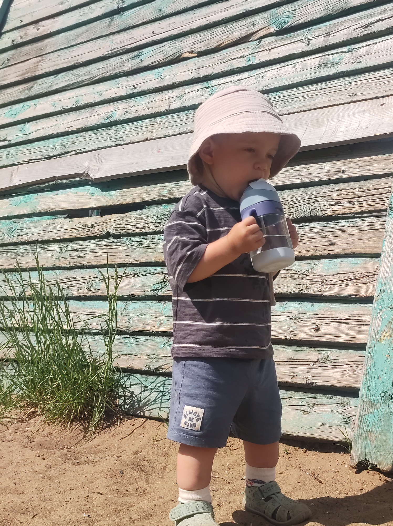 A young child stands in a sandy area in Chaikovsky, Russia, drinking from a blue and white water bottle. The child wears a beige bucket hat, a striped t-shirt, blue shorts with a "ALWAYS BE KIND" patch, white socks, and green sandals. They are standing in front of a weathered wooden wall with faded blue and green paint. Some grass grows to the left of the child. The ground is sand. The lighting suggests a sunny day.