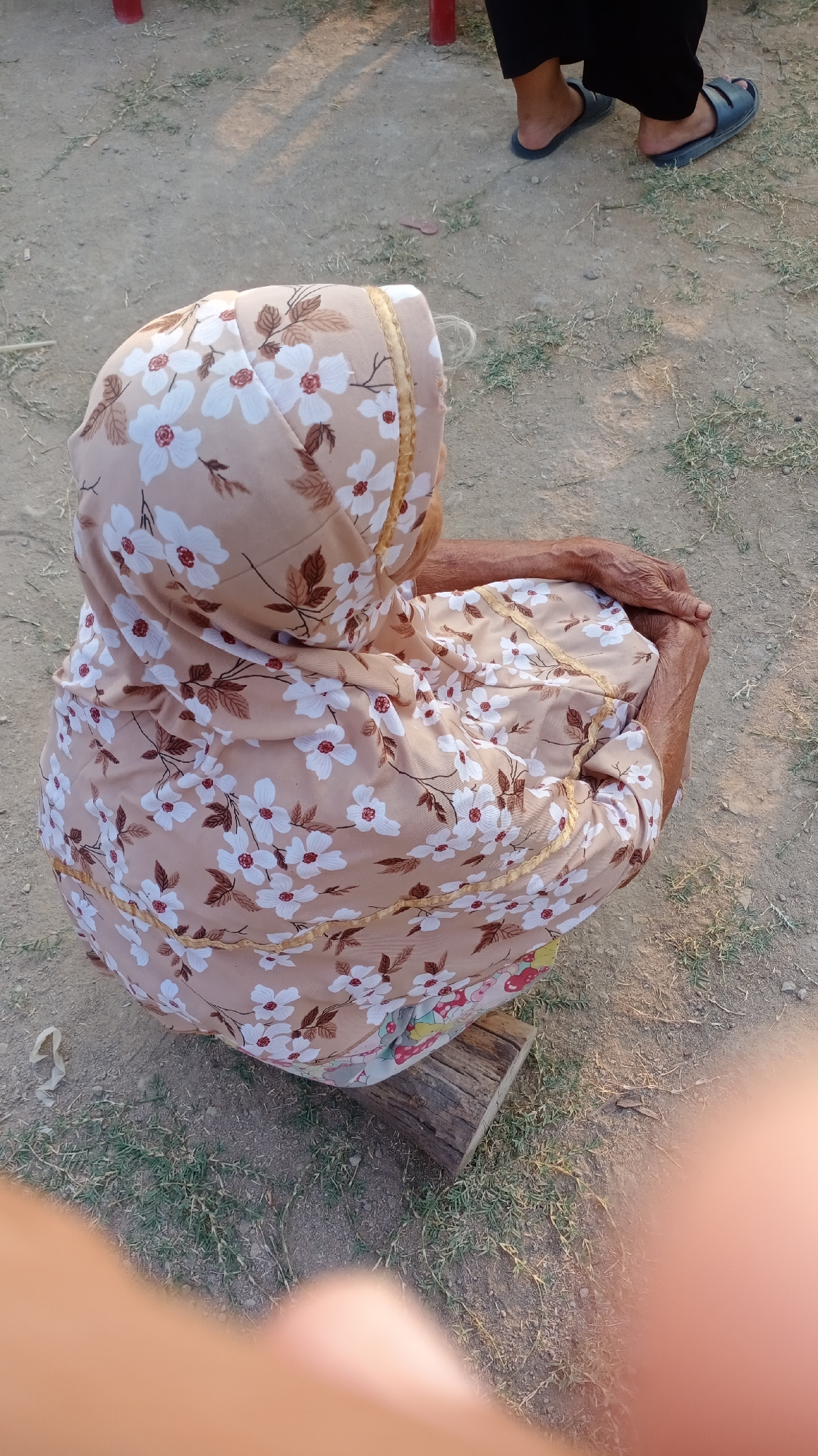 In Mertan, Indonesia, a person is shown from behind, crouching outdoors on a dirt and grass area. They are wearing a headscarf and a floral-patterned dress. Their hands are clasped in their lap. Next to them, a piece of wood serves as a makeshift seat. In the background, another person's legs and feet, clad in dark trousers and sandals, are visible. The scene appears to be a casual, outdoor gathering.