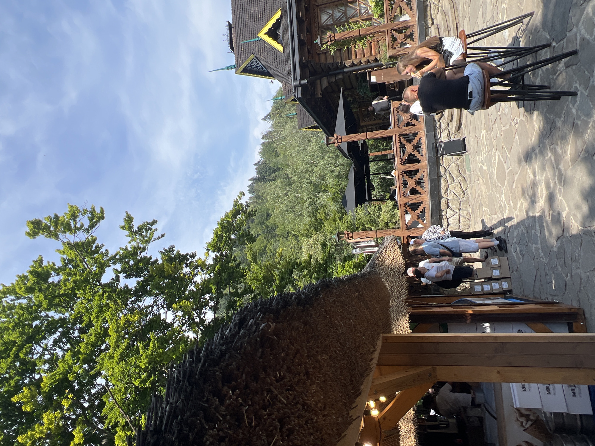 The image shows an outdoor seating area with a thatched roof stall on the left and a rustic wooden building on the right, likely a restaurant or cafe in Yaremche, Ukraine. The ground is paved with irregular stones. In the foreground, a man and a woman are seated at high stools at a small table. The man, wearing a black t-shirt and light blue jeans, is facing away from the camera. The woman, in a dark top and white skirt, is facing the man and holding a piece of food. Behind them, several other people are standing and walking around, some near the thatched roof stall where what appears to be ice cream is being served. The wooden building has a distinct, ornate roofline with yellow accents and dark shingles. Green trees and a hillside are visible in the background under a partly cloudy blue sky.