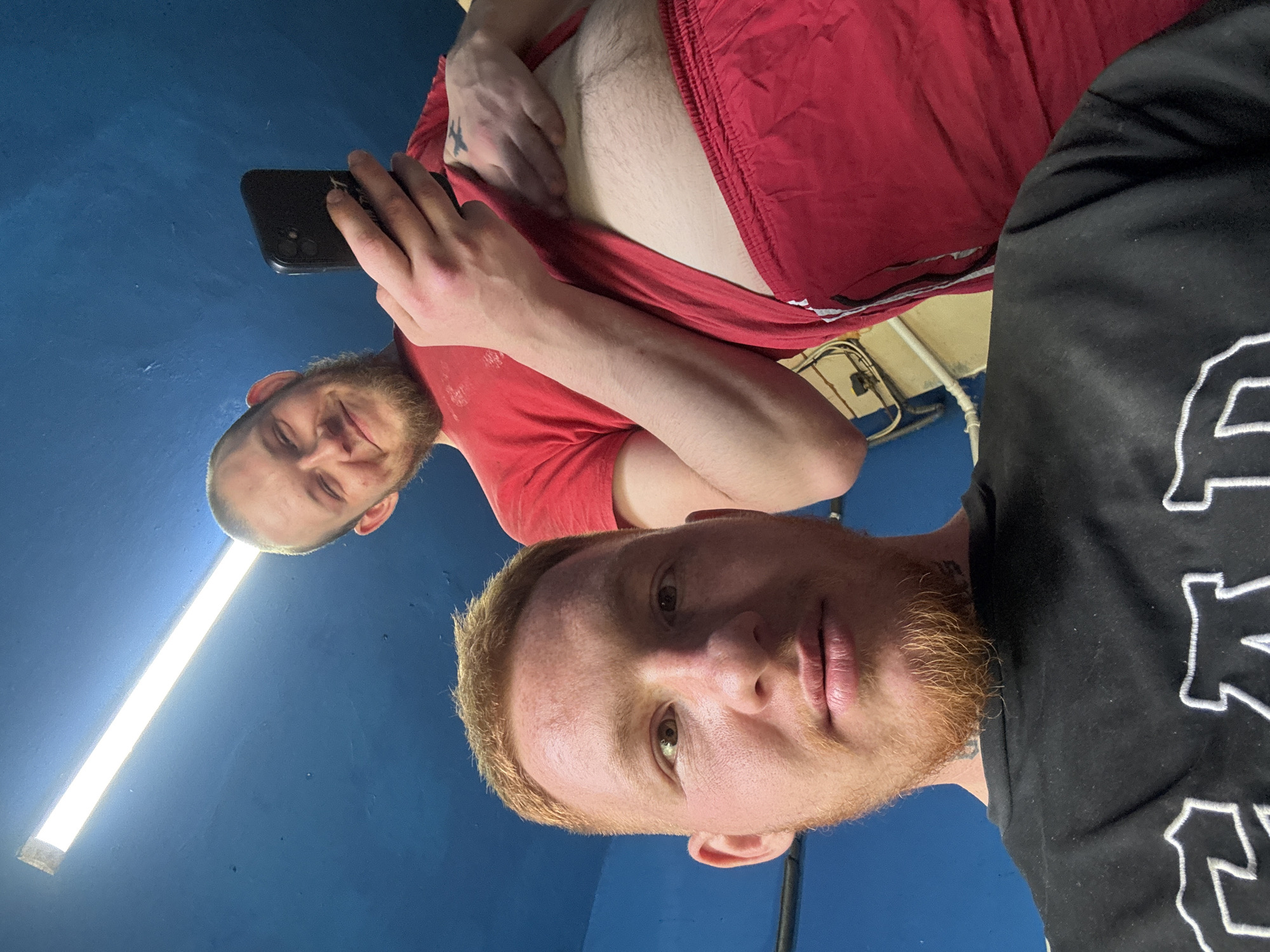 Two Caucasian males pose for a selfie in a room with a blue wall and a fluorescent light fixture on the ceiling. One male is wearing a black shirt with white lettering and has red hair and a beard. The other male is holding a cell phone and is wearing a red t-shirt and red shorts. His stomach is visible and he has a tattoo on his arm. Behind them is an electrical outlet and a metal pipe. The location of the photo is unknown but it is not believed to be in the city of Makiivka, Ukraine.
