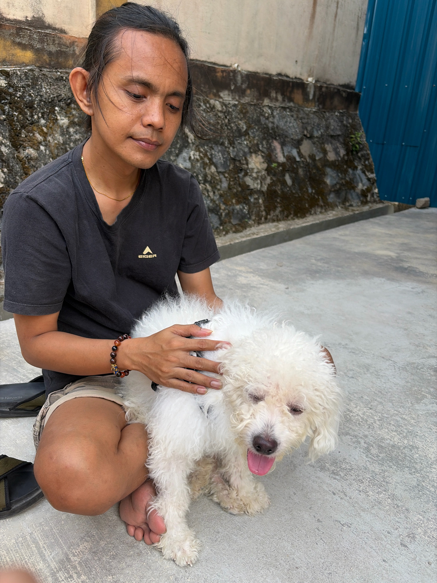 A person with dark, long, tied-back hair and dark clothing is sitting on the ground in front of a stone wall, petting a white, curly-haired dog. The person is wearing a dark t-shirt with a gold logo and patterned shorts. A beaded bracelet is visible on the person's right wrist. The dog is white with a pink tongue hanging out and appears to be panting. The ground is concrete, and a blue metal structure is visible in the background. The image was taken outdoors in Tanjung Pinang, Indonesia.