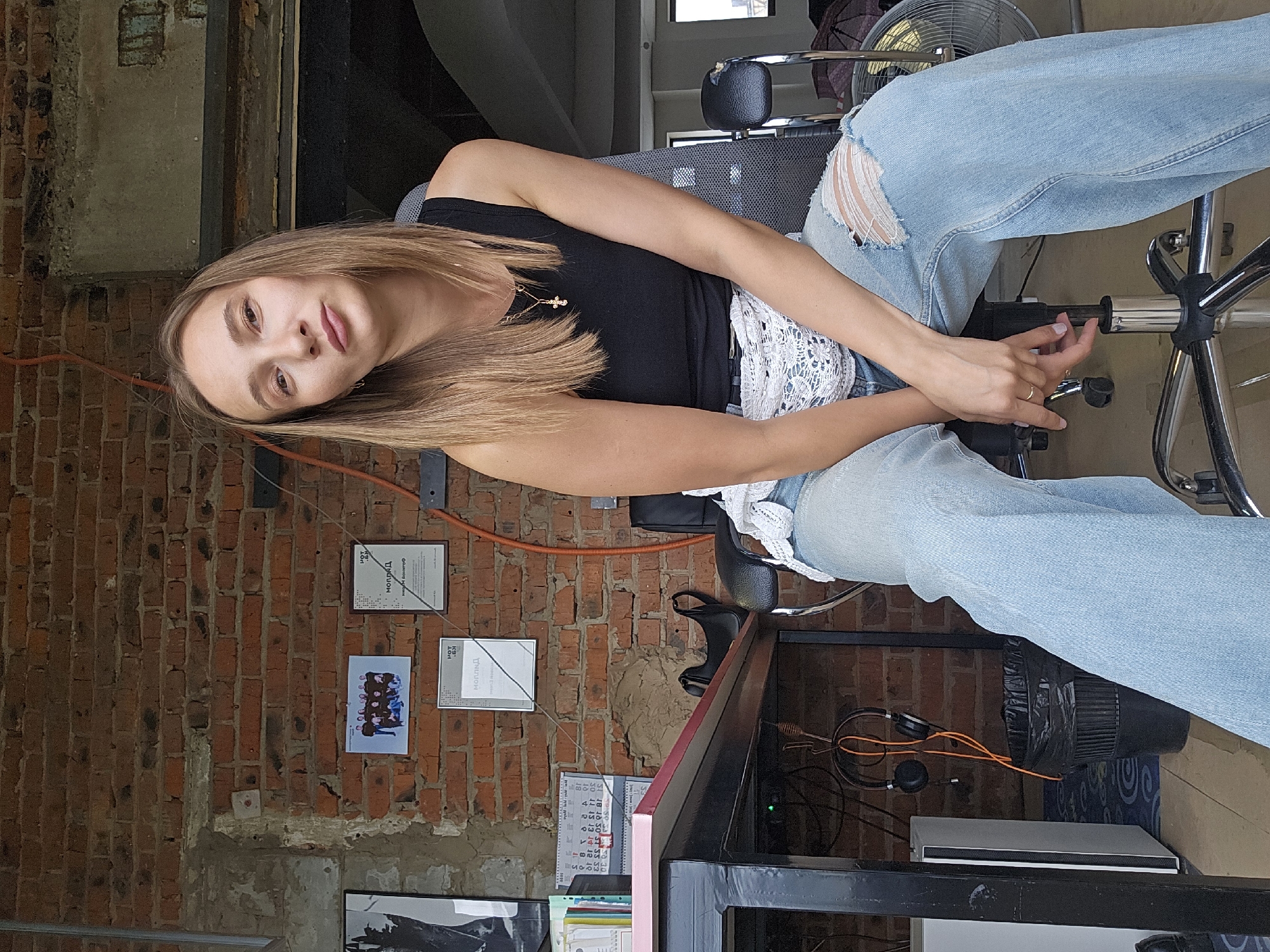 A woman is sitting in an office chair. She is wearing ripped jeans and a black tank top with a white lace belt. The background includes a brick wall with certificates and pictures hanging on it. There is a desk with a calendar and office supplies. Headphones are lying on the floor. The location is Chaikovsky, Russia.