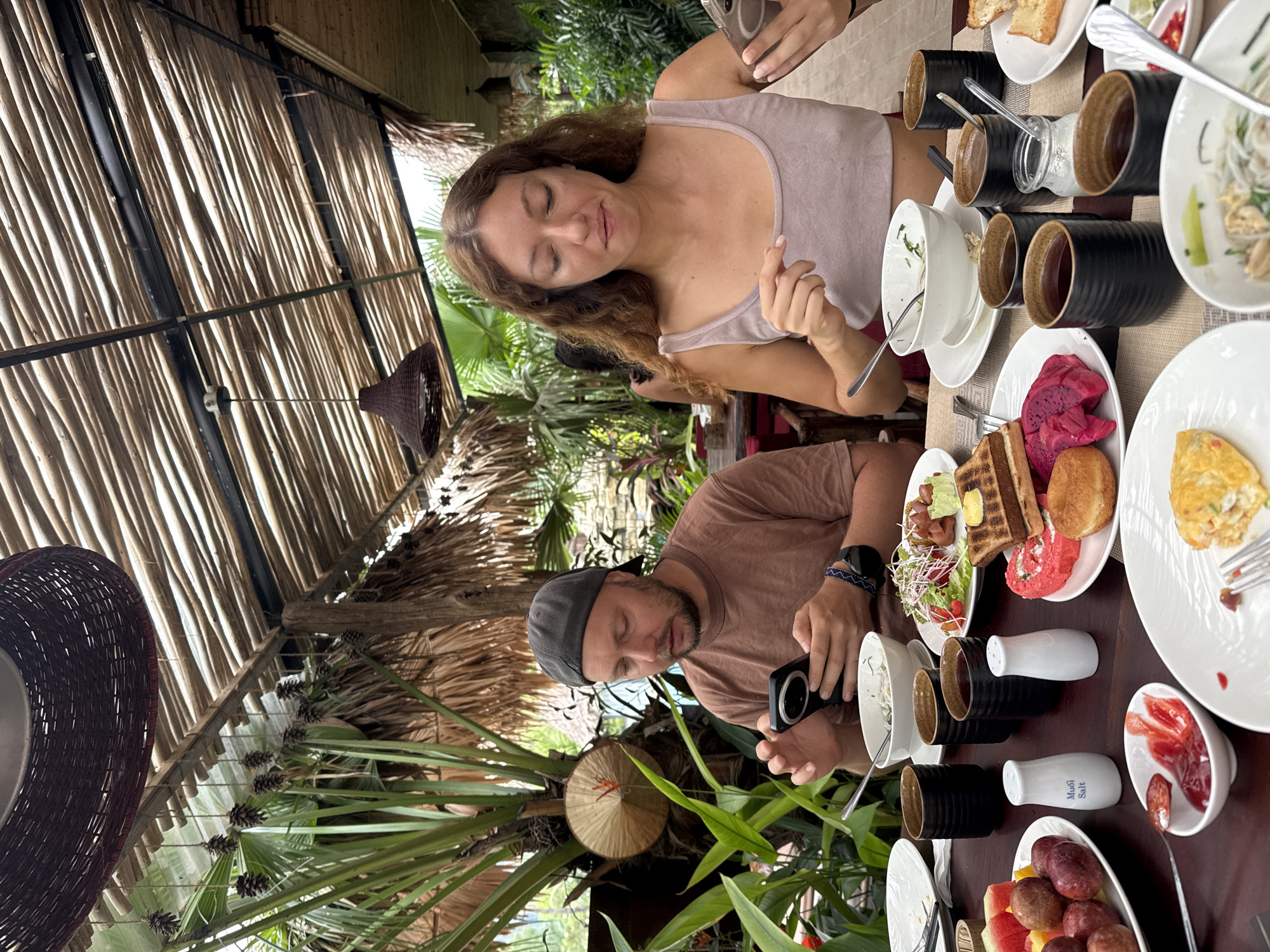 A man wearing a brown t-shirt and a grey baseball cap is seated at a wooden table, looking at his phone. A woman with long, curly brown hair, wearing a light purple tank top, is seated opposite him, also looking at her phone. Both are outdoors, under a thatched roof supported by wooden beams. In the background, lush green plants and palm trees are visible, suggesting a tropical environment. The table is laden with a breakfast spread, including bowls of food, cups of dark liquid, fruit, toast, and condiments. The overall setting appears to be a restaurant or an outdoor dining area in Nam Hoa Lư Ward, Vietnam, characterized by its natural and rustic ambiance.