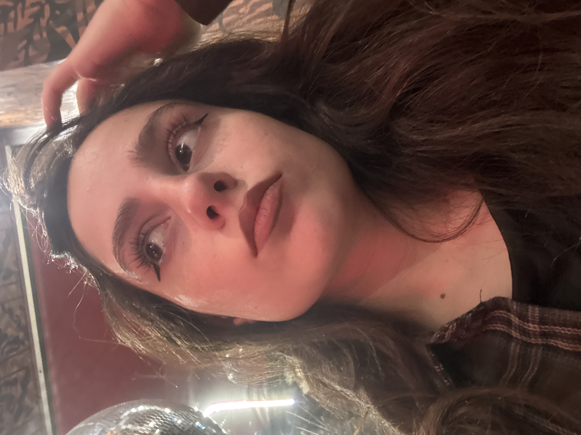 A close-up, low-angle shot features a female individual looking upwards and to her right. She has dark, long hair, styled with winged eyeliner and extended eyelashes. Her lips are full with a neutral-toned finish. Her right hand is visible, positioned near the upper right side of her head, appearing to touch or adjust her hair. She is wearing a dark garment, possibly a black top, with a segment of a brown and white plaid or striped pattern visible on her left shoulder, suggesting a jacket or shirt. The ambient lighting is dim with a prominent reddish hue, particularly on her face and neck.
To the upper left, a portion of a reflective disco ball is visible, along with a vertical, blurred light source that appears bright and possibly neon. The background features varied textures: a dark, patterned ceiling section above, a reddish-brown wall area, and a patterned, possibly upholstered, surface on the right side behind her head. The scene depicts an indoor entertainment venue, likely a bar or club, given the disco ball and ambient lighting. No specific visual identifiers within the image directly indicate a precise location in Yerevan, Armenia.