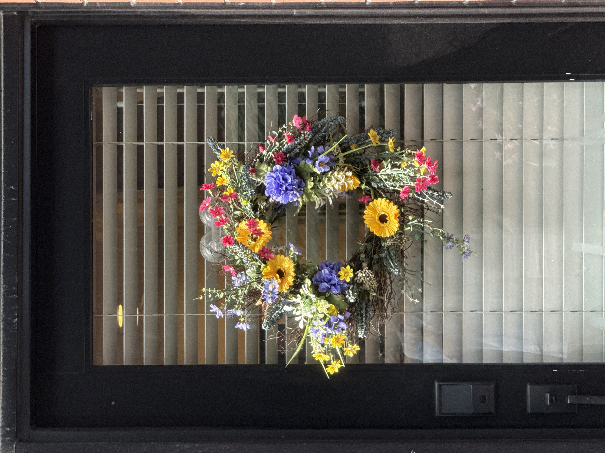 A floral wreath hangs on a glass-paned door, partially obscuring horizontal blinds. The wreath features a variety of artificial flowers in shades of yellow, pink, and blue, interspersed with green foliage and twigs. The door is framed by a dark material, likely metal, and a brick exterior is visible on the right side of the image. A modern black door lock is mounted on the left side of the door. The scene is set in Chicago, Illinois, United States of America.