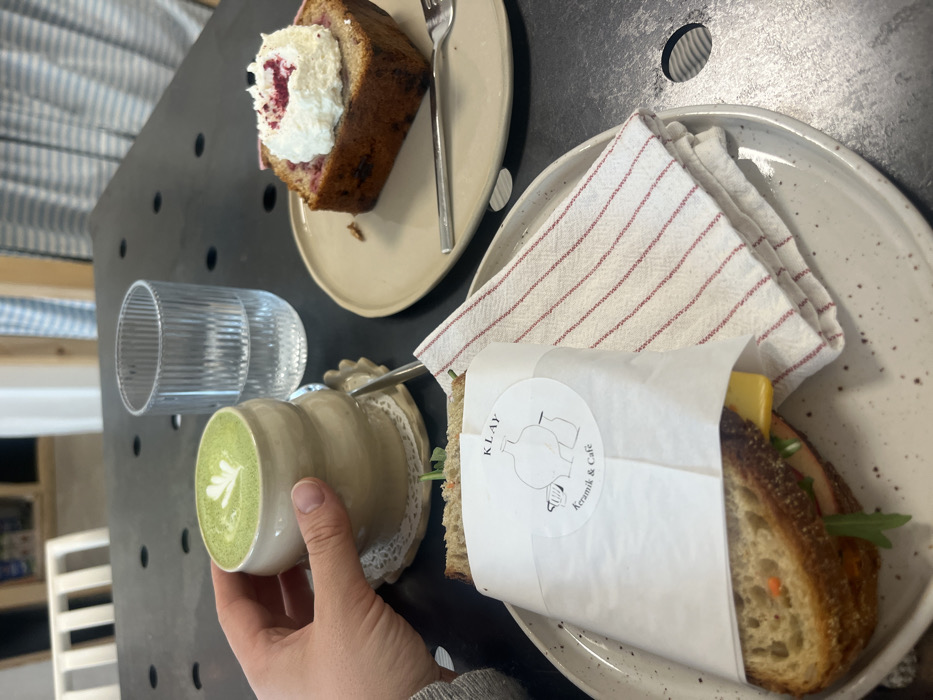 The scene depicts a cafe tabletop at KLAY Keramik & Café in Kaiserslautern, Germany. A light-skinned left hand grasps a beige, custom-shaped ceramic mug containing a green liquid with a white latte art design. This mug rests on a white, lace-patterned coaster. To its right, a clear, vertically ribbed glass holds water. On a light-colored, speckled ceramic plate, a sandwich wrapped in white paper with the "KLAY Keramik & Café" logo is visible, revealing dark bread, a slice of yellow cheese, and green and orange vegetable fillings. A folded white cloth napkin with thin red stripes lies beside the sandwich. On an adjacent, plain light-colored ceramic plate, a rectangular slice of baked goods, topped with white whipped cream and red sprinkles, is presented with a silver fork. The table surface is dark gray with circular perforations. The background is out of focus, showing blurred light-colored wooden furniture and a light blue and white striped textile.