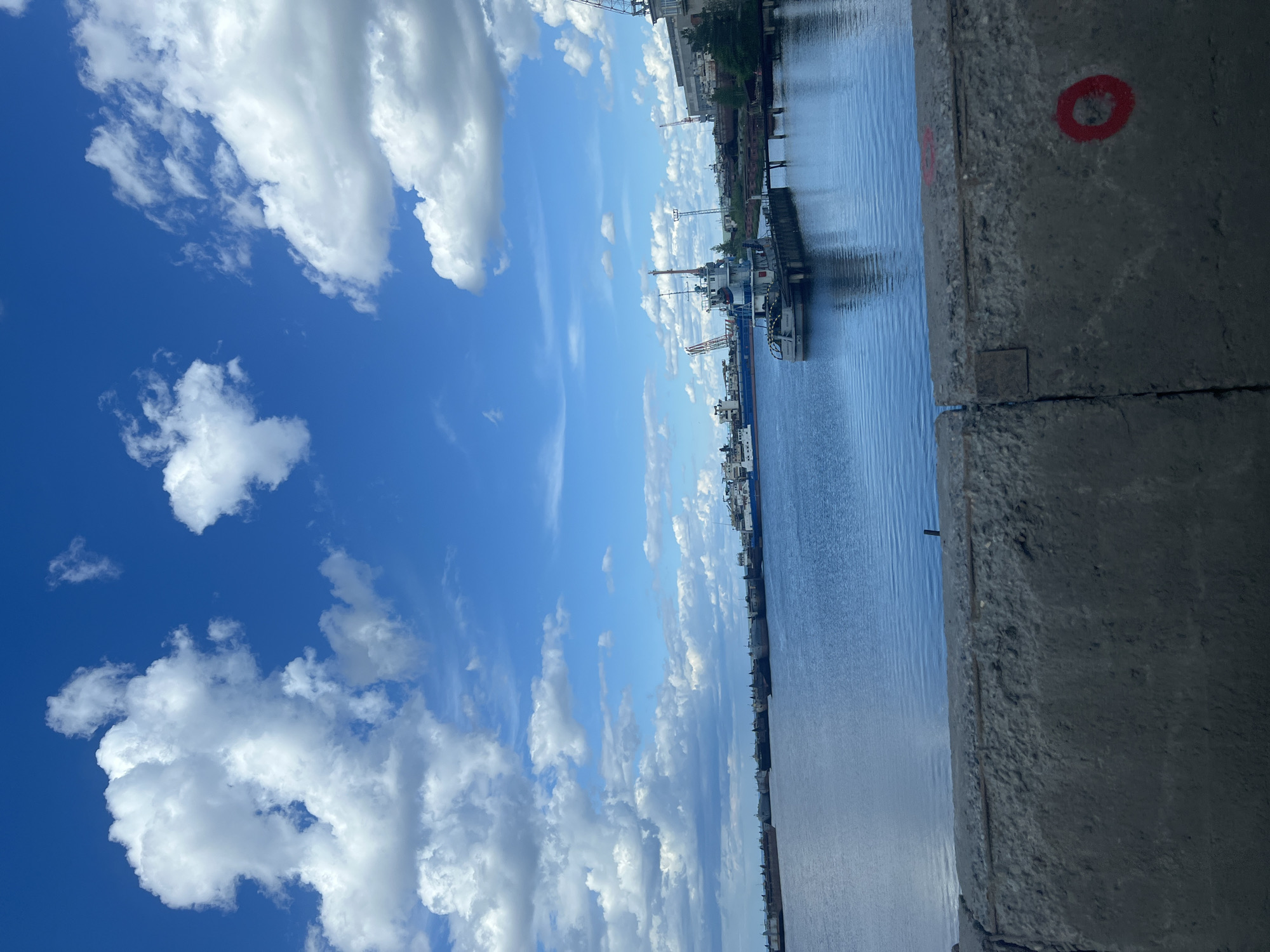 The image presents a view from an elevated concrete structure, possibly a bridge, in the city of Chaikovsky, Russia. The scene encompasses a body of water, likely a river or a wide canal, with several vessels docked along the shoreline. The sky is blue with scattered cumulus clouds. The concrete structure in the foreground appears worn, with a red circle painted on the surface. Buildings are visible along the distant shoreline.