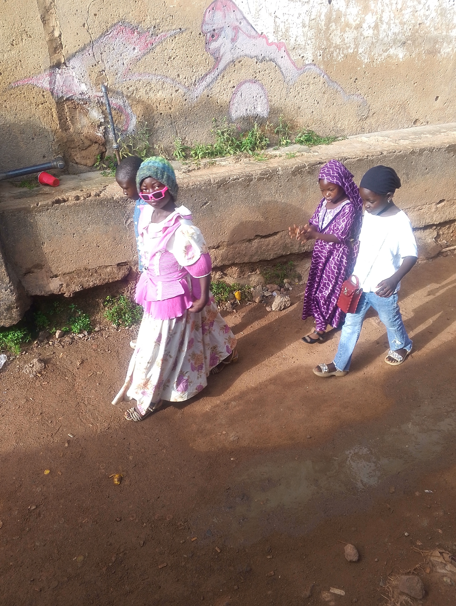 The image depicts four children walking on a dirt path next to a concrete wall. The wall has some graffiti and plants growing along its base. The path appears to be in an urban area, possibly on the outskirts of a town or city, given the unpaved ground and the wall. The first child, at the front of the group, is wearing a long floral skirt, a pink and white top, a knitted hat and pink sunglasses. The second child, partly obscured, wears blue clothing. The third child is wearing a purple printed dress and a headscarf of the same fabric. The fourth child is wearing jeans, a white t-shirt, a black head covering and has a small red purse. It cannot be confirmed that this image is from Jos, Nigeria.