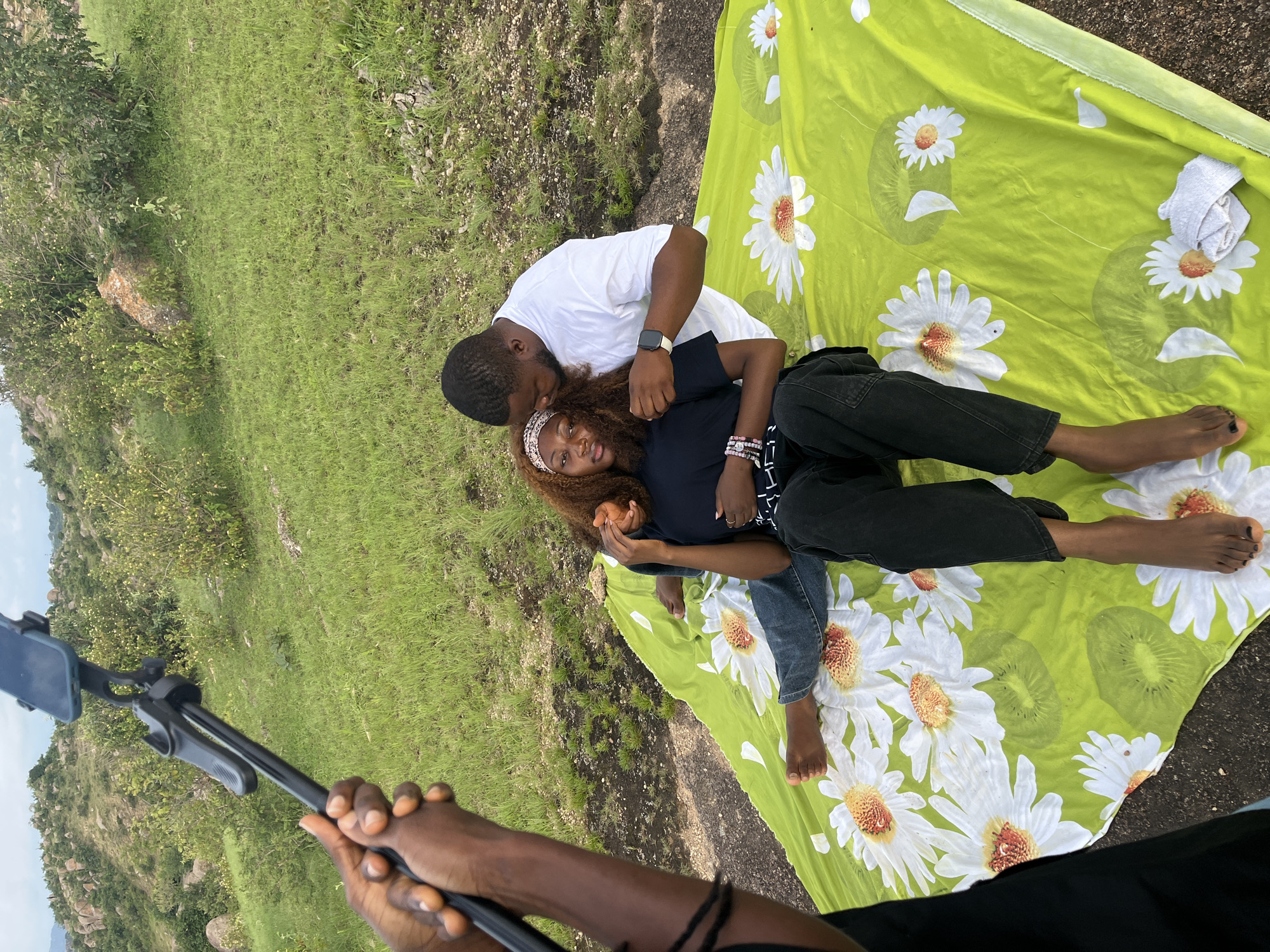 In a grassy, hilly outdoor location, a person is holding a selfie stick with a phone attached, aiming it towards a couple sitting on a bright green blanket adorned with white flowers and kiwi slices. The woman, seated, is wearing a black t-shirt and dark pants, with a patterned headband in her auburn hair. She holds an unidentified object in her hand. The man, also seated and turned towards her, is wearing a white t-shirt and has his arm around her shoulder, whispering in her ear. The background consists of green grass, scattered rocks, and vegetation.