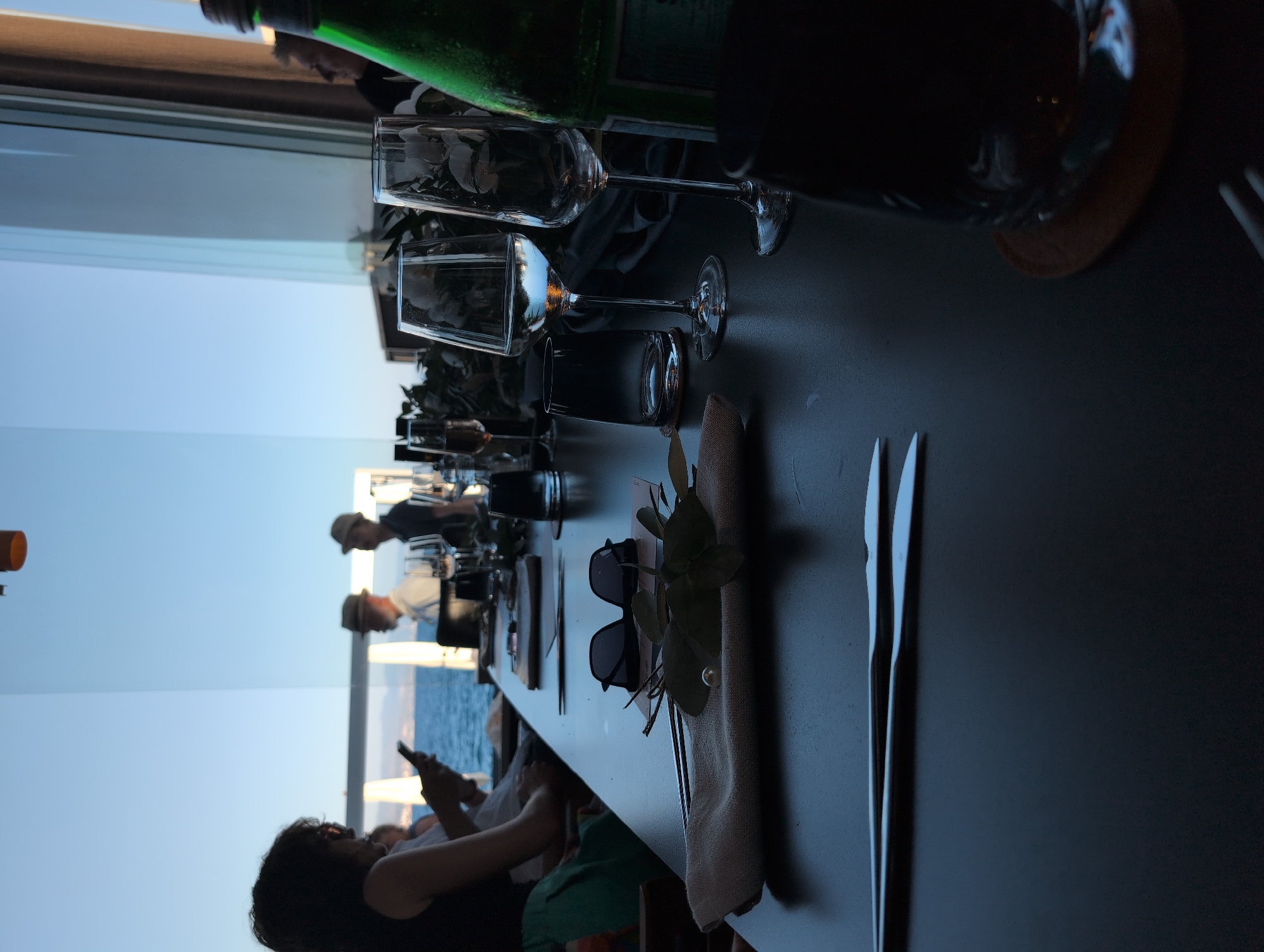 A group of individuals are seated at a long, dark table in a restaurant with a large window overlooking the sea and a clear blue sky. The setting appears to be a fine dining establishment, with formal place settings including silverware, wine glasses, and napkins. In the foreground, a pair of sunglasses rests on a napkin adorned with a pearl and green leaves. Further down the table, wine glasses and a bottle of green liquid are visible. Several people are partially visible, some engaged with their phones, others in conversation. The overall atmosphere is serene and sophisticated, with the natural light from the sea creating a relaxed ambiance. The location is the coastal city of Opatija, Croatia.