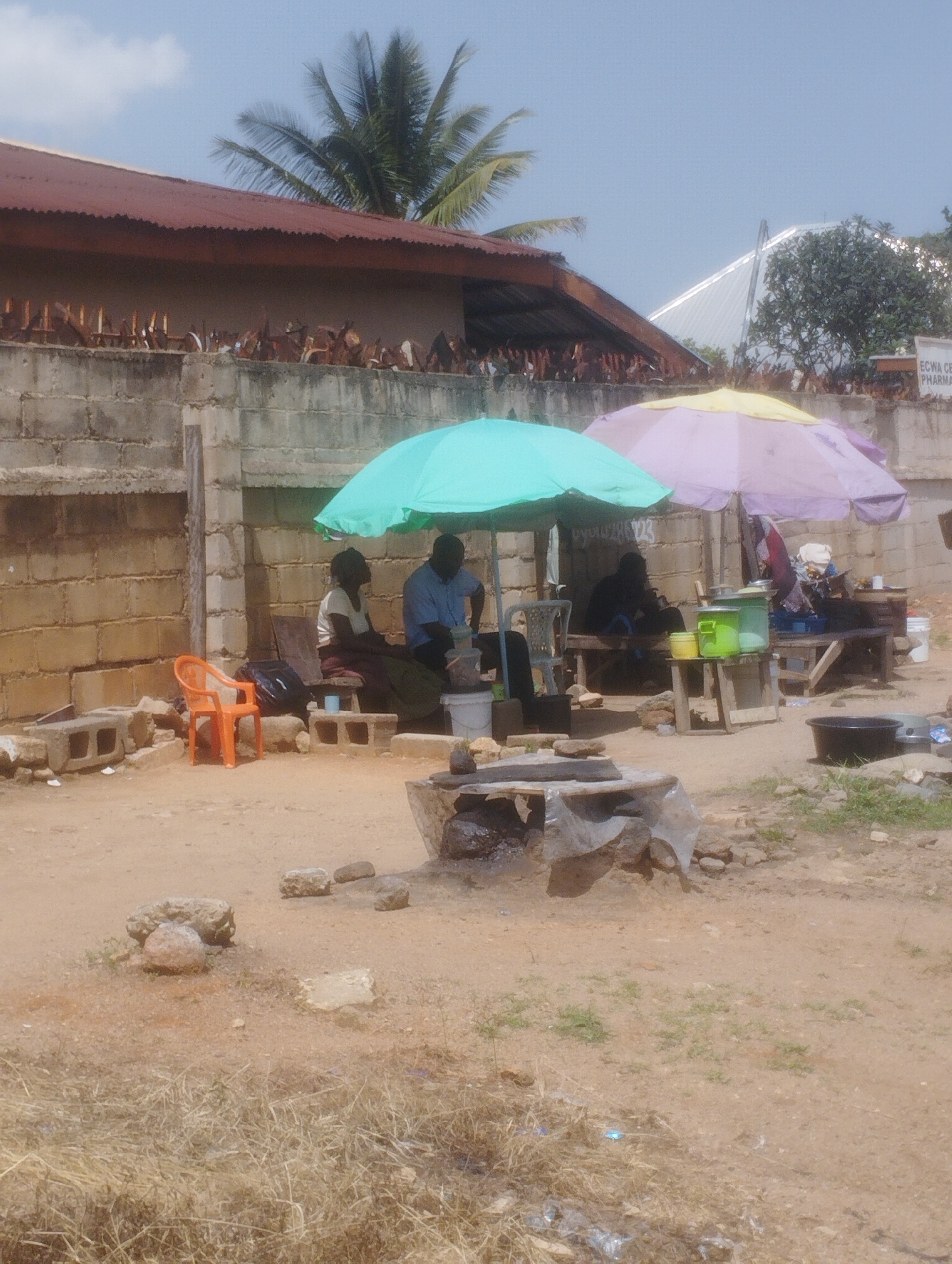 The image depicts an outdoor scene in Jos, Nigeria, likely a market or roadside vending area. In the foreground, dry, dusty ground is visible with scattered rocks and dead vegetation. A makeshift cooking area constructed from stones and possibly cement is in the center-foreground.
Further back, a man in a blue shirt and a woman in a white top and patterned skirt are seated under a turquoise umbrella. They appear to be interacting with items on a table between them, which includes containers and possibly food or drink. To the right of this setup, another umbrella, this one pink and yellow, shades a different seating area where a person in a dark top is seated. Several wooden benches and tables are arranged, with various items like containers and what appear to be cloths or garments visible.
Behind the seating areas, a wall constructed from concrete blocks forms a backdrop. Above this wall, the edge of a building with a reddish-brown, corrugated metal roof is visible, along with a tall palm tree. In the distance, a modern bridge structure with a white, arched design is prominent against a light blue sky with scattered clouds. A sign with faint lettering, possibly indicating "ECWA CENT PHARMAC," is also visible on the wall. The overall impression is of a casual, functional outdoor commercial space.