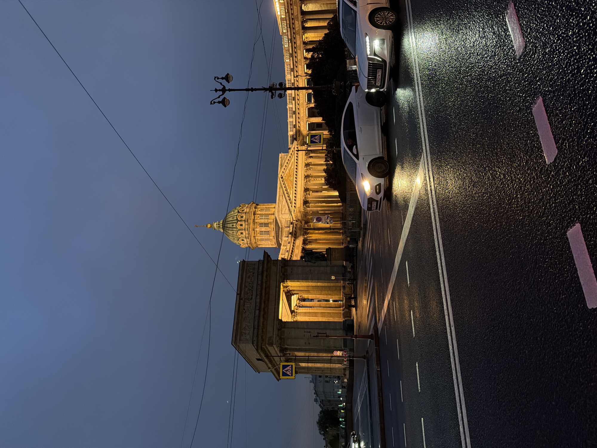 This is a full, eye-level, outdoor shot of Kazan Cathedral in Saint Petersburg, Russia, taken at dusk. The cathedral is illuminated and appears to be the focal point of the scene. A colonnade extends from the main structure. In front of the cathedral, a neoclassical archway with columns is also lit. Two white sedans are driving on a wet, asphalt road in the foreground, their headlights illuminating the road. The sky is a dark, twilight blue. There is a pedestrian crossing in front of the archway. A lamppost with multiple lamps is visible on the right side of the image. The street is wet, suggesting recent rain, and reflections of the lights can be seen on the surface.