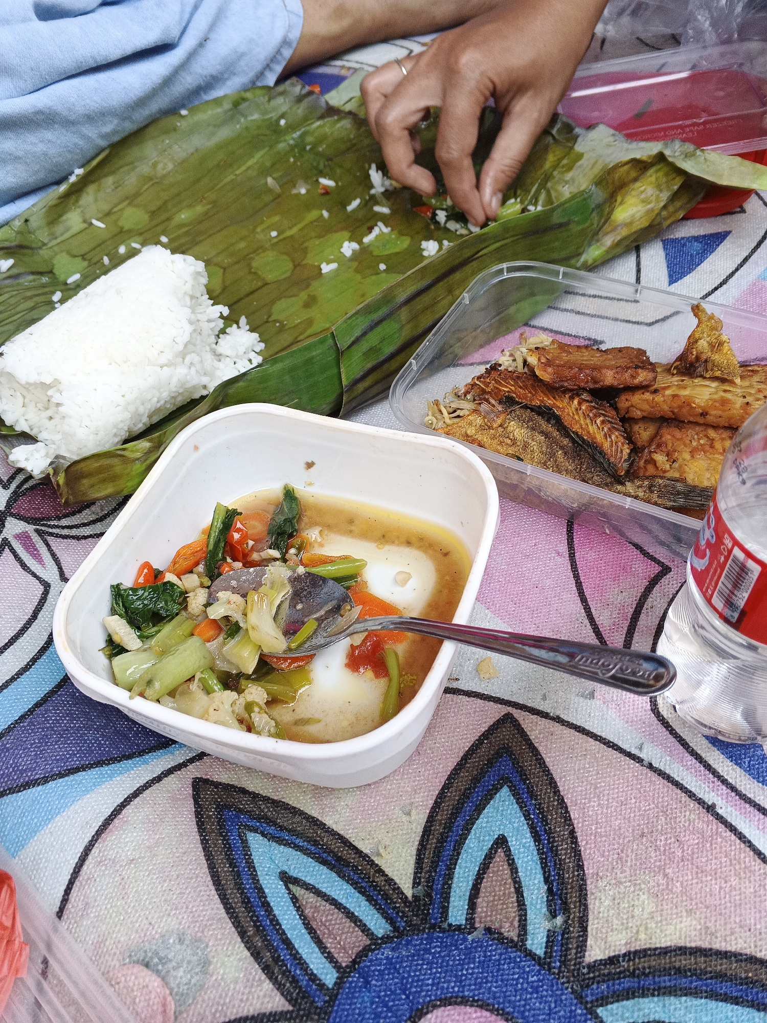 In Warungkiara, Indonesia, a person's hands are visible as they arrange rice wrapped in a banana leaf. Nearby, a white bowl contains a stew with vegetables such as bok choy, carrots, cauliflower, and tomatoes. Adjacent to the bowl is a clear plastic container filled with fried fish and tempeh. A spoon rests in the stew, and a bottle of water is partially visible on the right. The scene is set on a colorful tablecloth with a floral pattern.