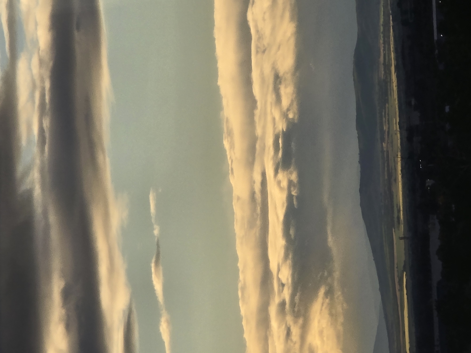 The image depicts a landscape at dusk or dawn. The sky is filled with streaks of clouds, ranging from white to grey, illuminated by the setting or rising sun. Below the sky is a low-lying landmass, possibly hills or mountains, leading to a flat expanse of land. On the right side of the image, the environment transitions into darkness, possibly indicating the edge of the city or a wooded area. Some light sources, possibly streetlights or building lights, are visible in the dark region.