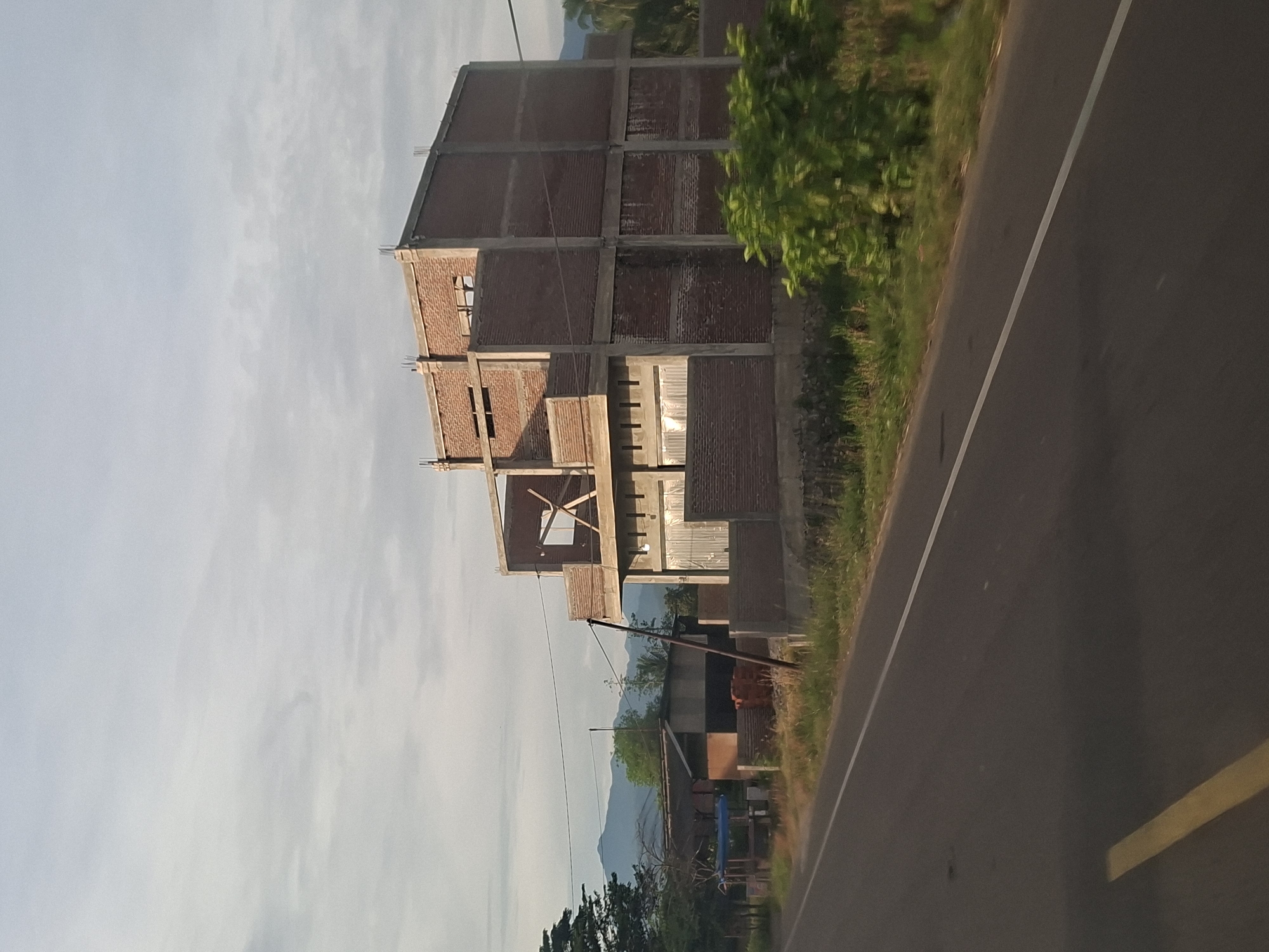 A long shot captures a three-story building under construction in Meukokuthang, Indonesia. The building, made of red brick and concrete, has an unfinished facade with exposed rebar at the top. To the left of the building are smaller structures, one with a corrugated metal roof and another with a blue tarp. A paved road with white and yellow lane markings runs diagonally across the foreground. In the background, a lush green landscape leads to distant mountains under a hazy sky.