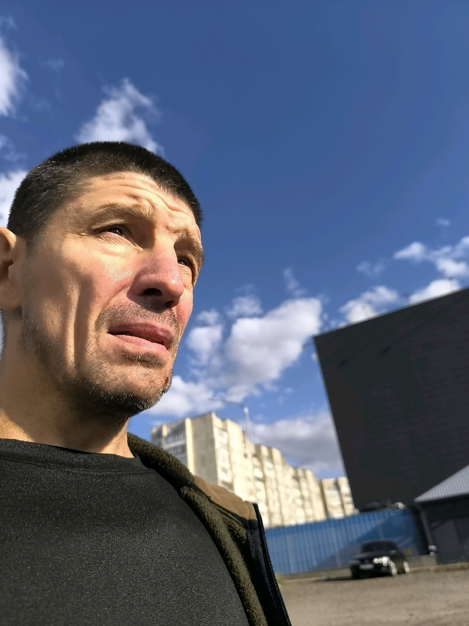 The image shows a close-up, low-angle shot of a man's face and upper body, set against a bright blue sky with scattered white clouds. The man has short dark hair, a weathered complexion, and is wearing a dark t-shirt and a green jacket. His gaze is directed upwards and to the right, with a slightly furrowed brow. In the background, out of focus, are several buildings, including a light-colored multi-story structure and a dark, angular building. A blue fence and a dark car are also visible further in the background. The overall impression is of an outdoor urban setting, possibly in Qarağandı city administration, Kazakhstan, on a sunny day.