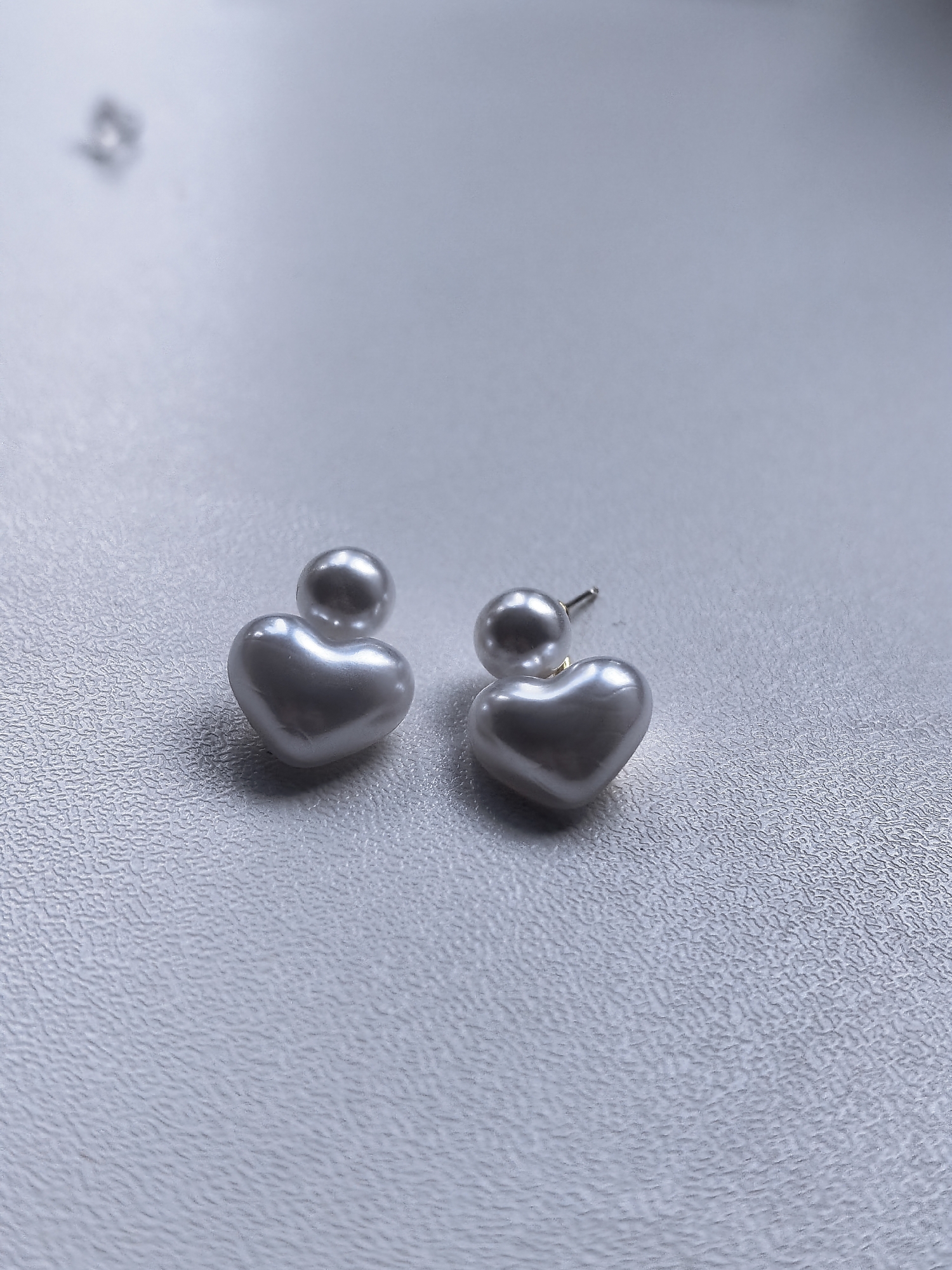 A pair of pearl earrings, each designed with a heart-shaped pearl below a smaller spherical pearl, are displayed on a textured, light-colored surface. One earring is positioned on the left side of the frame, while the other is to its right. The lighting casts subtle shadows, highlighting the smooth, lustrous quality of the pearls. The background is soft and out of focus, drawing attention to the jewelry. There is no indication of people, actions, interactions, or the environment in Sterlitamak, Russia.