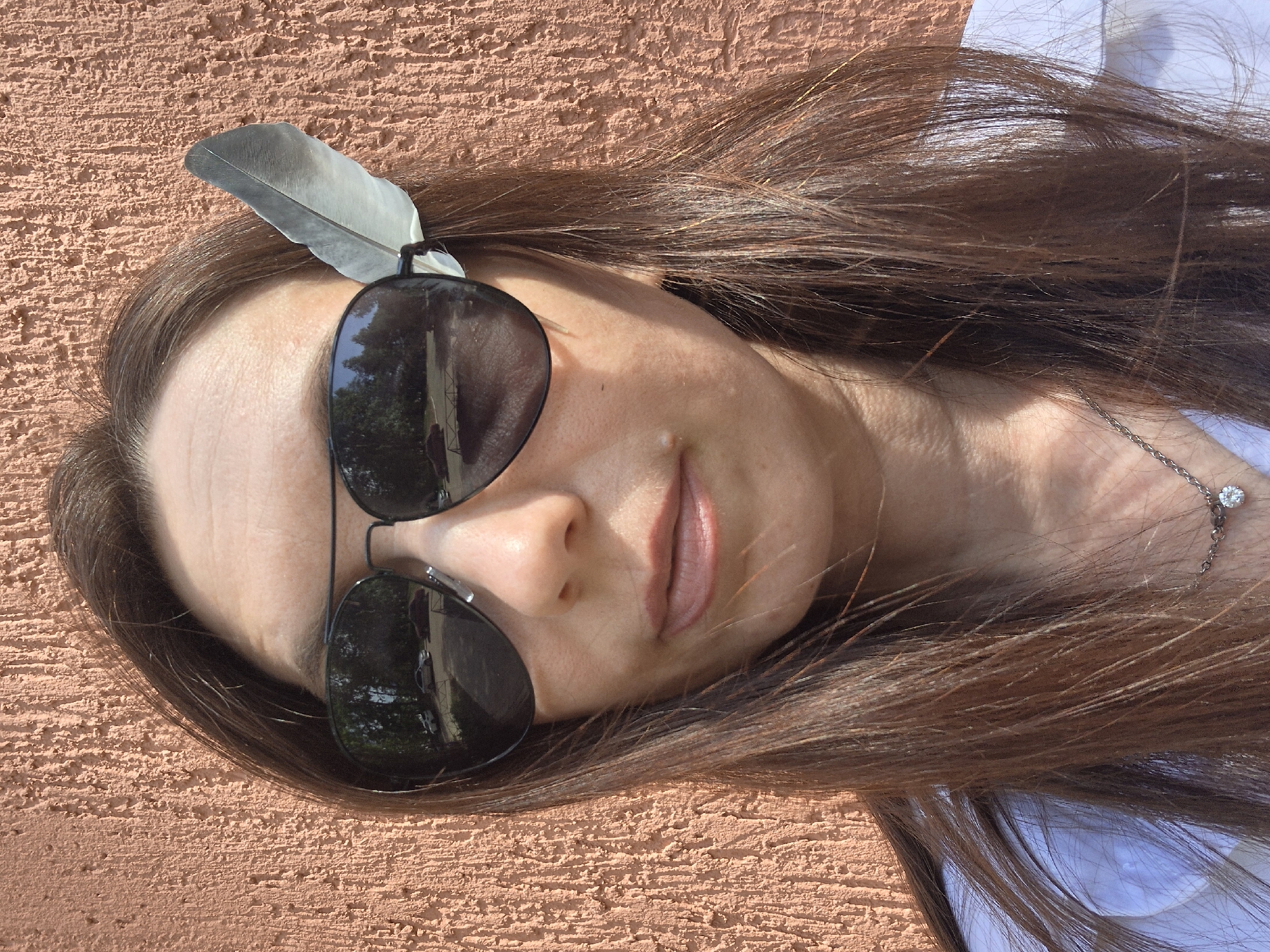 The image shows a woman lying on a light red textured surface, possibly a wall. She is wearing sunglasses and a feather adorns her hair near the glasses. She has brown hair and appears to be wearing a white shirt. A delicate silver necklace with a small diamond pendant is visible around her neck. The image appears to have been taken outdoors, with the sunglasses reflecting trees and buildings, although the location is unidentifiable.