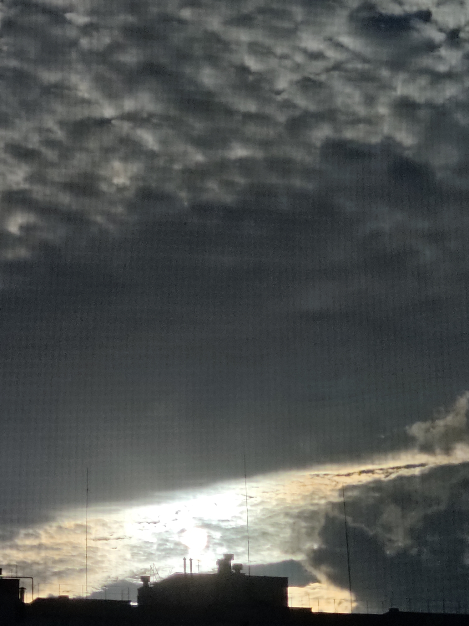 A silhouette of rooftops and communication towers in Odesa, Ukraine, is visible against a dramatic sky filled with dark, textured clouds. A bright band of sunlight breaks through the clouds, creating a stark contrast and illuminating the undersides of some of the cloud formations. The scene appears to be captured during either sunrise or sunset.