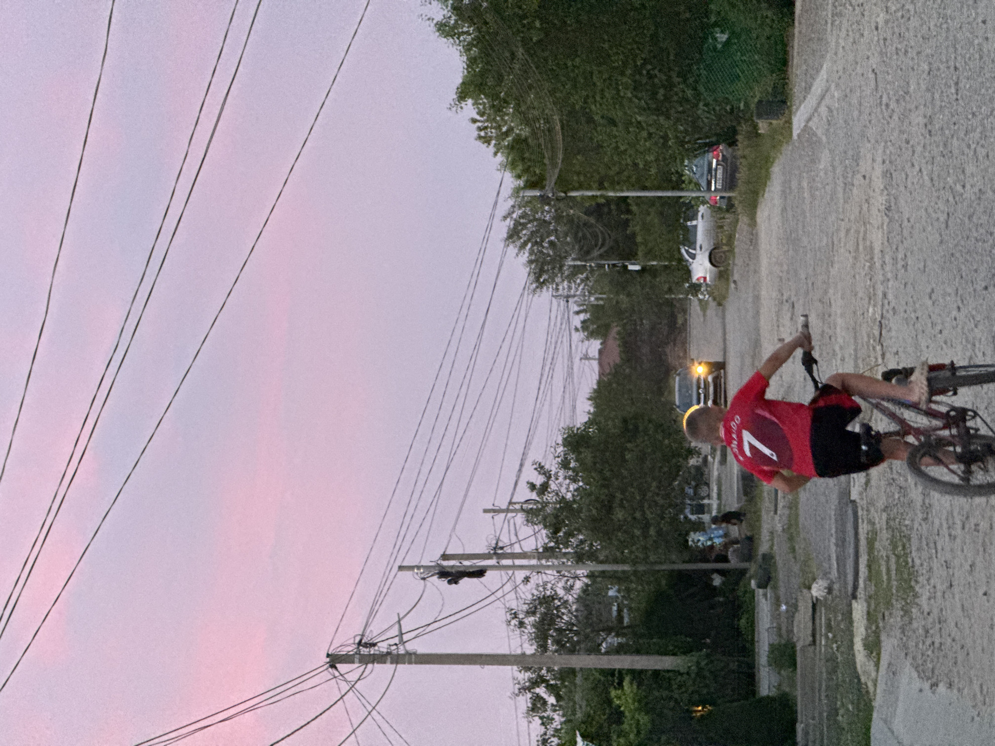 In Mikhaylovsk, Russia, at dusk, a young boy wearing a red jersey with the number 7 and black shorts rides a bicycle down a gravel road. Overhead, numerous power lines stretch across the pink and purple sky. To the left, utility poles stand tall, supporting the tangled network of wires, with lush green trees and bushes flanking the road. In the background, several cars are visible. One car with its headlights on approaches from further down the road, while two other cars are parked on the right side. The environment appears to be a residential area with a slightly unpaved road.
