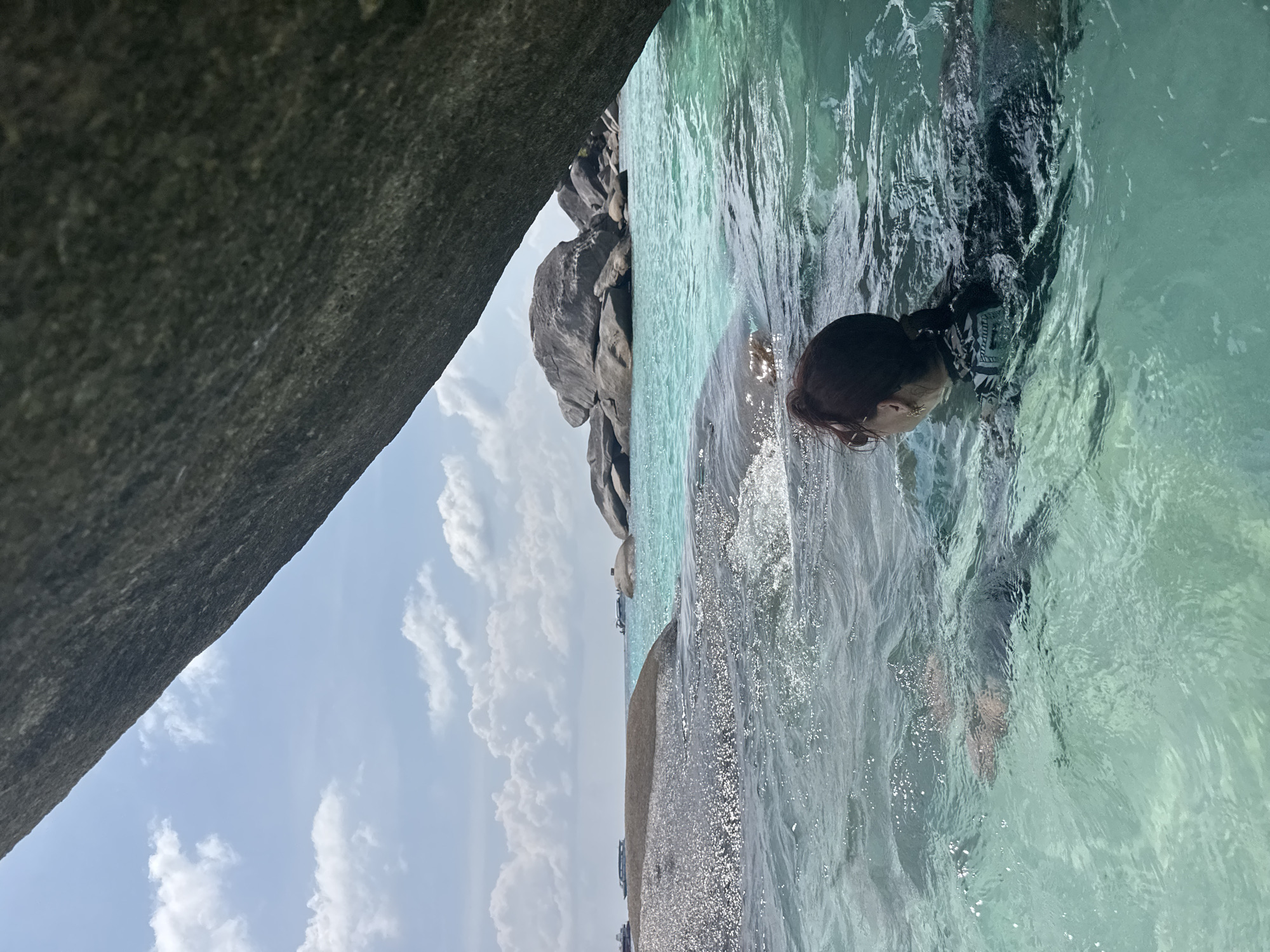 A woman with dark hair pulled back in a bun swims in clear, turquoise water near large rocks. The location appears to be coastal with rocky outcroppings and a partly cloudy sky above. Based on the location indicated, the scene is likely near the coast of Lam Kaen, Thailand, where similar geological features are common.
