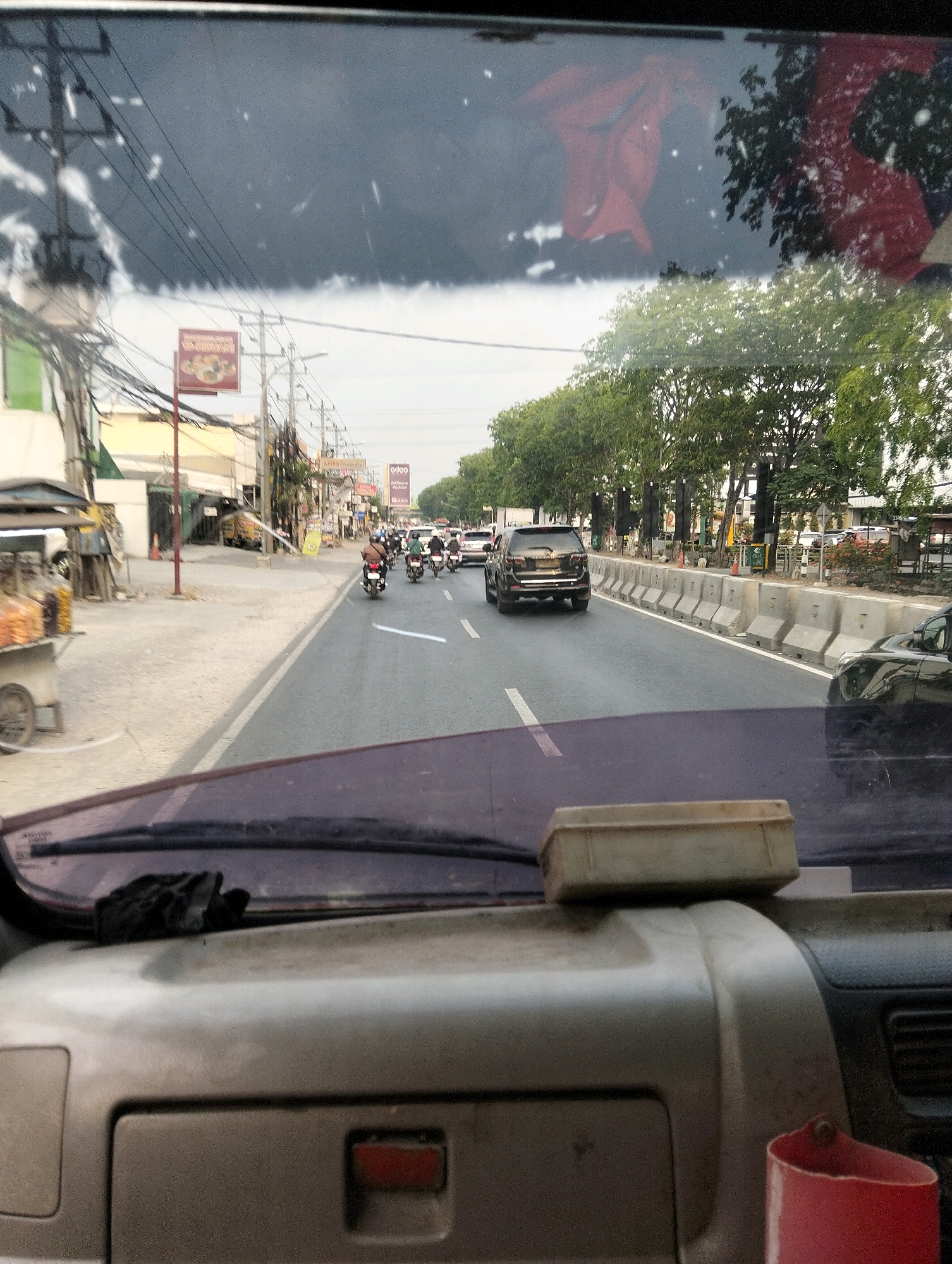 The image is taken from the interior of a vehicle, looking forward through the windshield. The scene depicts a street in Semarang, Indonesia, with a moderate amount of traffic. On the left side of the road, there are buildings and a food cart. The road itself has multiple lanes and is bordered by concrete barriers and trees on the right. Several motorcycles and cars are visible on the road, with most riders wearing helmets. A black SUV is prominent in the middle lane. The sky appears overcast. In the foreground, the dashboard of the vehicle is visible, with a small rectangular object placed on top of it. A red cup is partially visible in the bottom right corner. Reflections of trees and red objects are present on the windshield.
