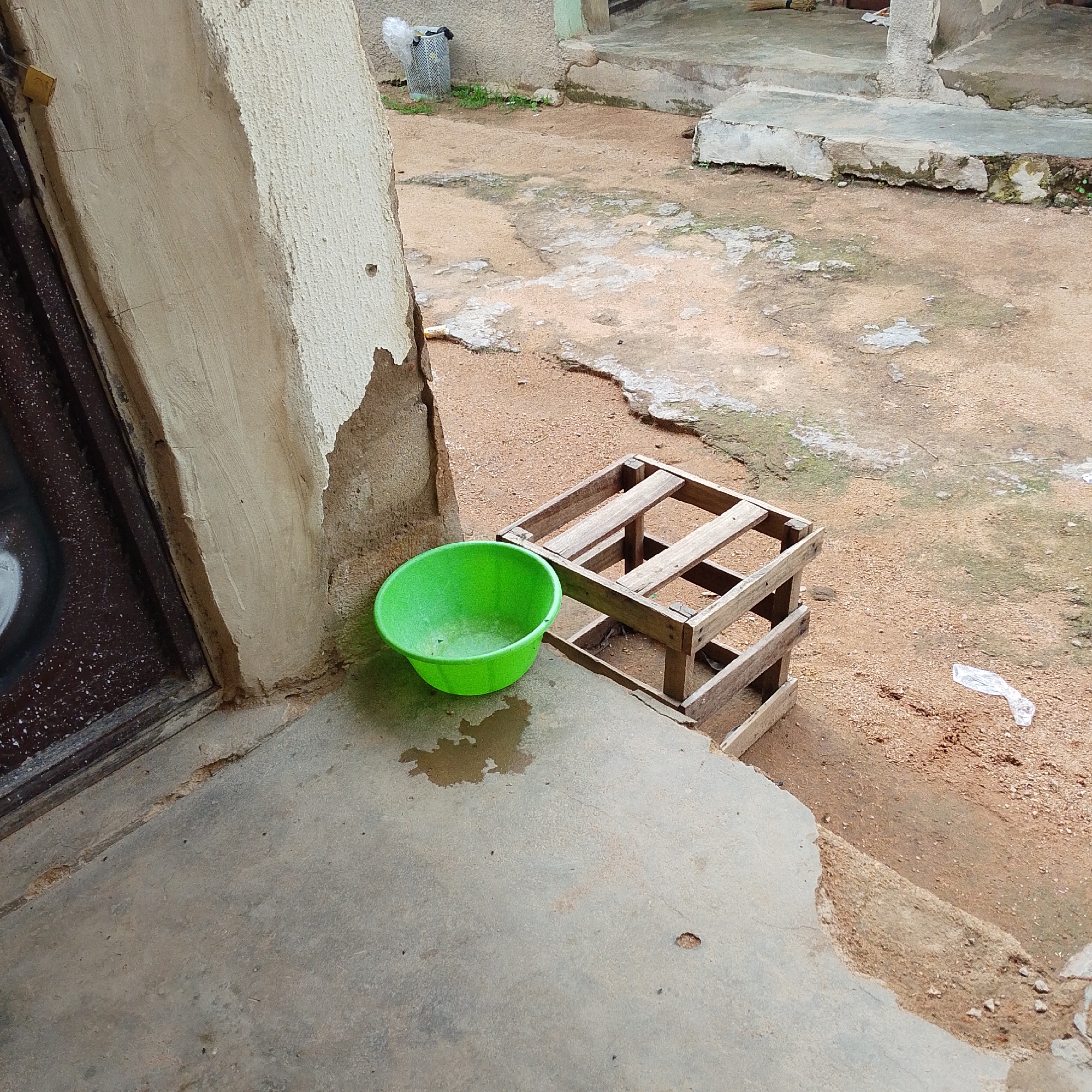 This image depicts an outdoor scene in the city of Jalingo, Nigeria, featuring a weathered concrete entryway, a bright green plastic basin, and a crude wooden crate. The entryway, positioned on the left, shows signs of disrepair with peeling plaster and a dark, possibly wooden doorframe. The concrete ground in front of the entryway is wet, with a visible puddle beneath the green basin. The basin itself is round and appears to be empty. To the right of the basin is a disassembled wooden crate, constructed from rough-hewn planks. The ground surrounding these objects is a mix of dirt and gravel, with some patches of dried mud or lighter-colored earth. In the background, a concrete structure with a weathered texture is visible, along with a grey wall and hints of greenery. A crumpled piece of white plastic is lying on the ground to the right of the wooden crate. The lighting suggests it is daytime, with diffused sunlight.
