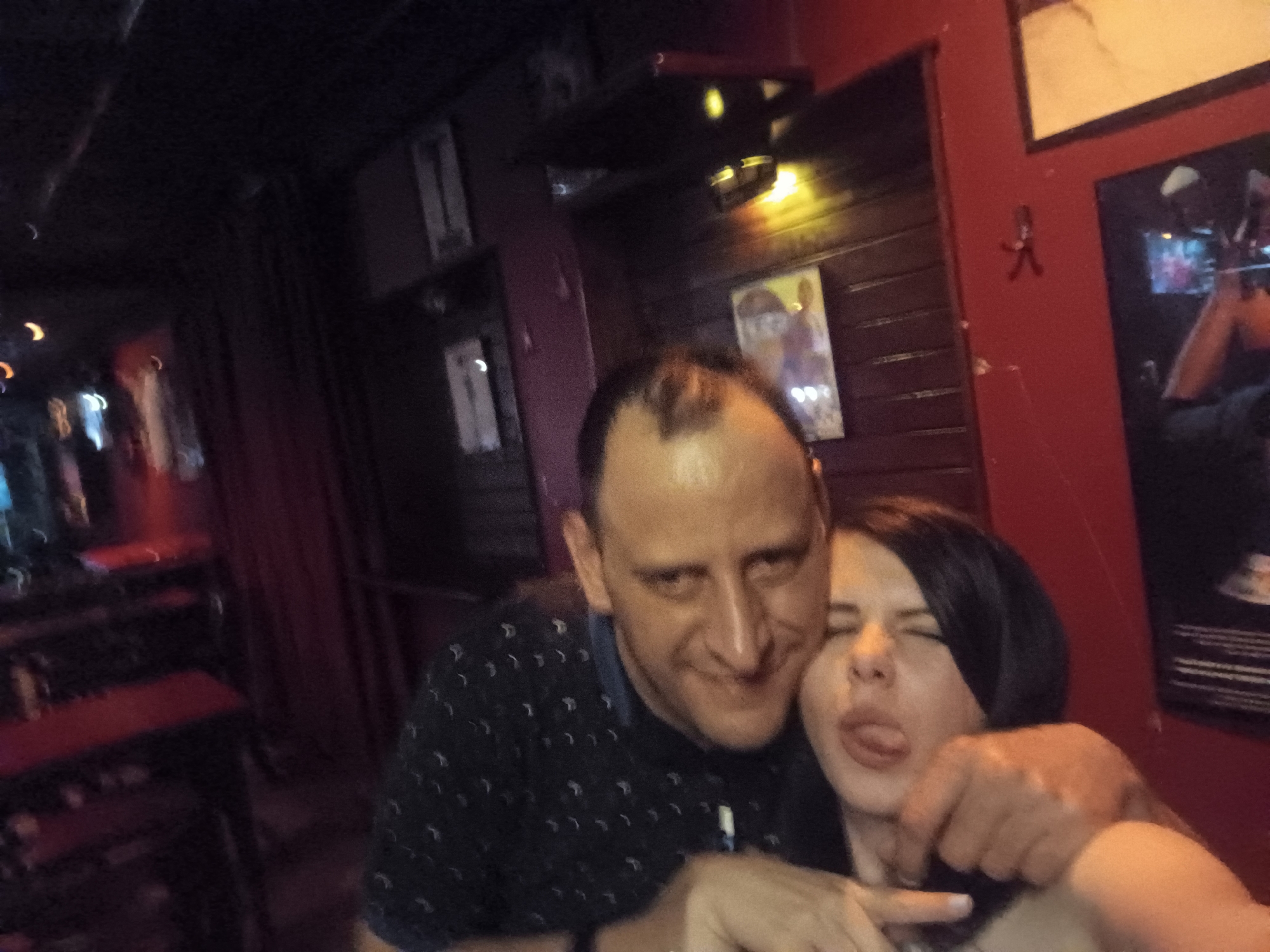 The image shows a man and a woman posing for a selfie in a dimly lit interior. The man, fair-skinned with short hair, is wearing a dark shirt with a light pattern. He has his arm around the woman. The woman, fair-skinned with dark hair, is sticking her tongue out. The background includes wooden walls, framed pictures on the wall, and red curtains. A dimly lit bar with tables and stools is visible in the background. The location is likely a bar or restaurant in Sofia, Bulgaria, based on visual cues.