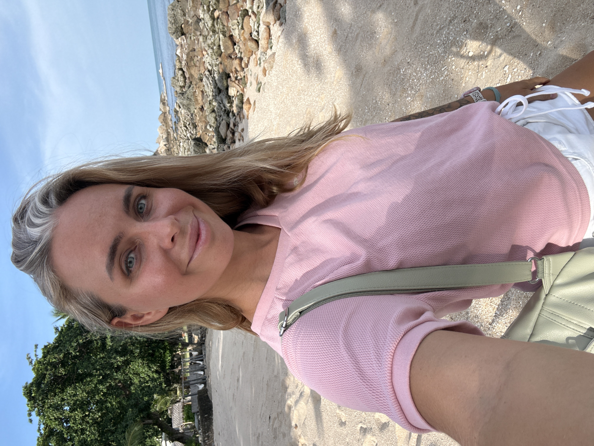 A woman is taking a selfie on a beach in Ko Samui District, Thailand. The woman has blonde hair with gray streaks and blue eyes, and she is smiling at the camera. She is wearing a pink t-shirt and white shorts. A light green purse is slung over her shoulder. The background of the image shows a sandy beach, rocky coastline, and the ocean under a clear blue sky. To the left, there is a large tree and some buildings with thatched roofs.