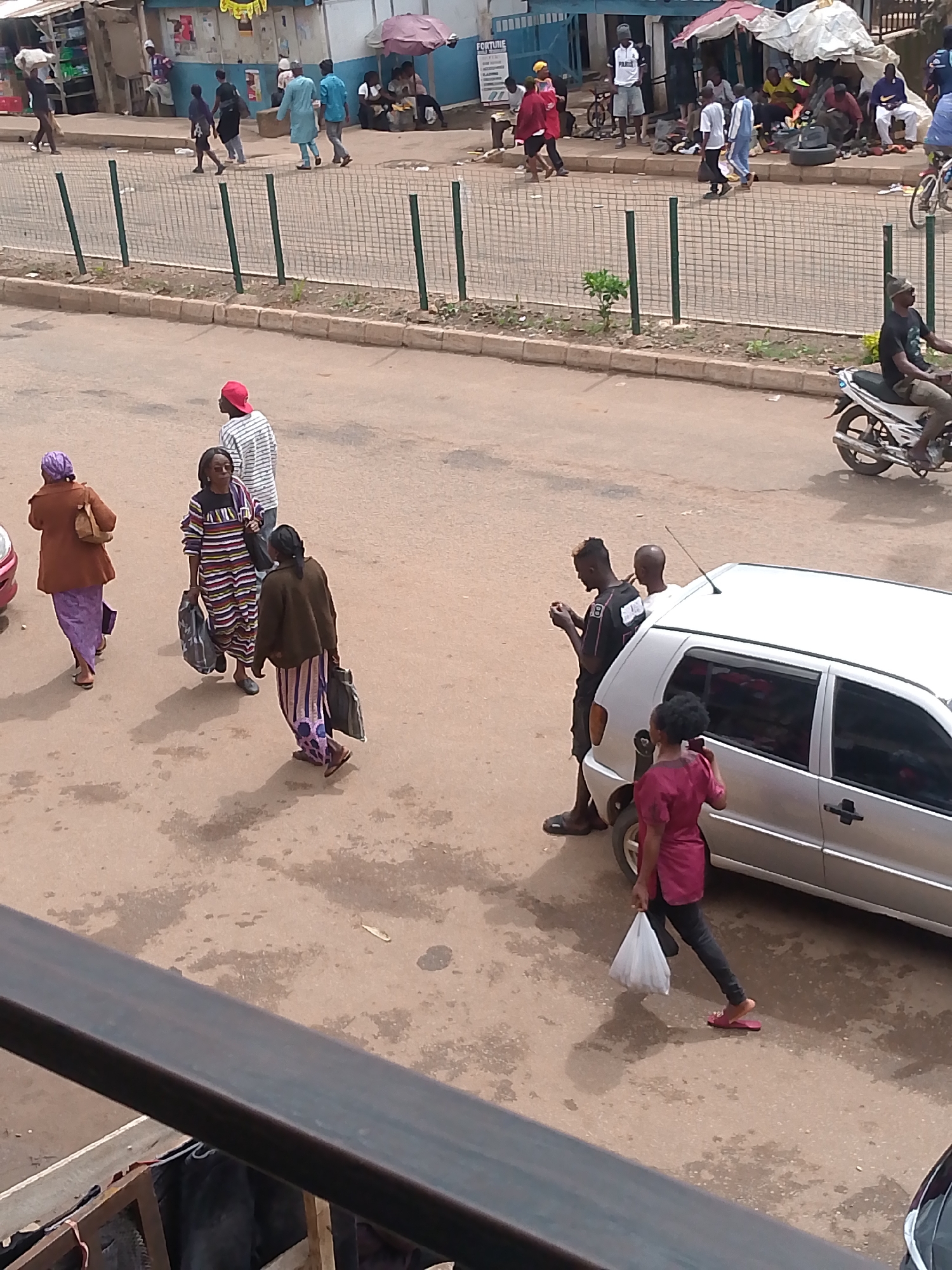 A street scene in Bukuru, Nigeria, features a light-colored car parked on a dirt road. Three women are walking on the road; one in a brown jacket and purple dress carries a brown purse, another in a striped dress carries two black bags, and a third in a dark top and striped pants carries a black bag. A man wearing a red cap and striped shirt walks away from the camera. A man in a black t-shirt is on his phone near the car. A woman in a burgundy outfit is walking away from the car with a white plastic bag. In the background, a white motorcycle with a rider is on the road. Other pedestrians and market stalls are visible further down the street.