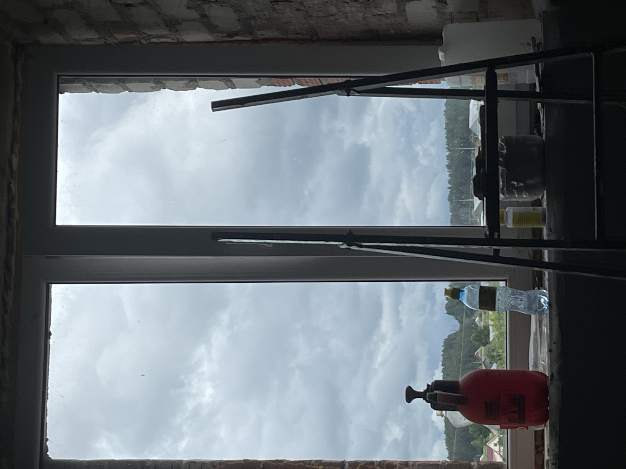 The image shows an indoor scene looking out of a double-paned window. The window frame is grey. The lower section of the window has a view of a cloudy sky and some trees and buildings in the distance, suggesting an urban or semi-rural environment. A red sprayer bottle and a water bottle are positioned on the windowsill. In the foreground, a dark, possibly metal, structure partially obstructs the view. The brick surrounding the window suggests an unfinished or partially renovated construction project. The location is Фоки, Russia.