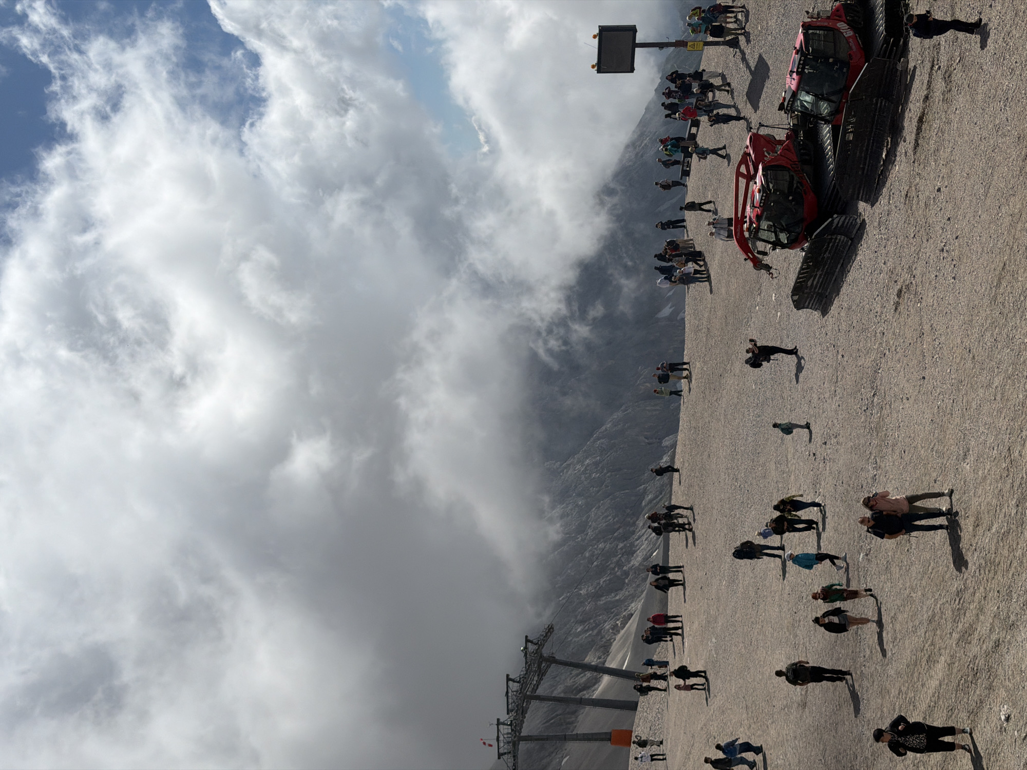 The image depicts a high-altitude scene with a wide, gravelly plateau populated by numerous tourists. In the background, imposing mountains are partially obscured by dramatic, swirling clouds. A ski lift structure, comprising metal towers and cables, is visible on the left side of the frame, indicating a mountainous recreational area. Two large, red snow groomers are parked prominently in the foreground, suggesting the area may be used for winter sports. A digital display board is mounted on a pole to the right. The people, dressed in casual attire, are scattered across the plateau, some walking, others standing in small groups, and a few are taking photographs. The ground is covered in small stones and gravel, with patches of snow or ice visible in the distance on the mountain slopes. The sky is a mixture of blue patches and dense, overcast clouds. The scene appears to be a viewpoint or a station at a significant elevation.