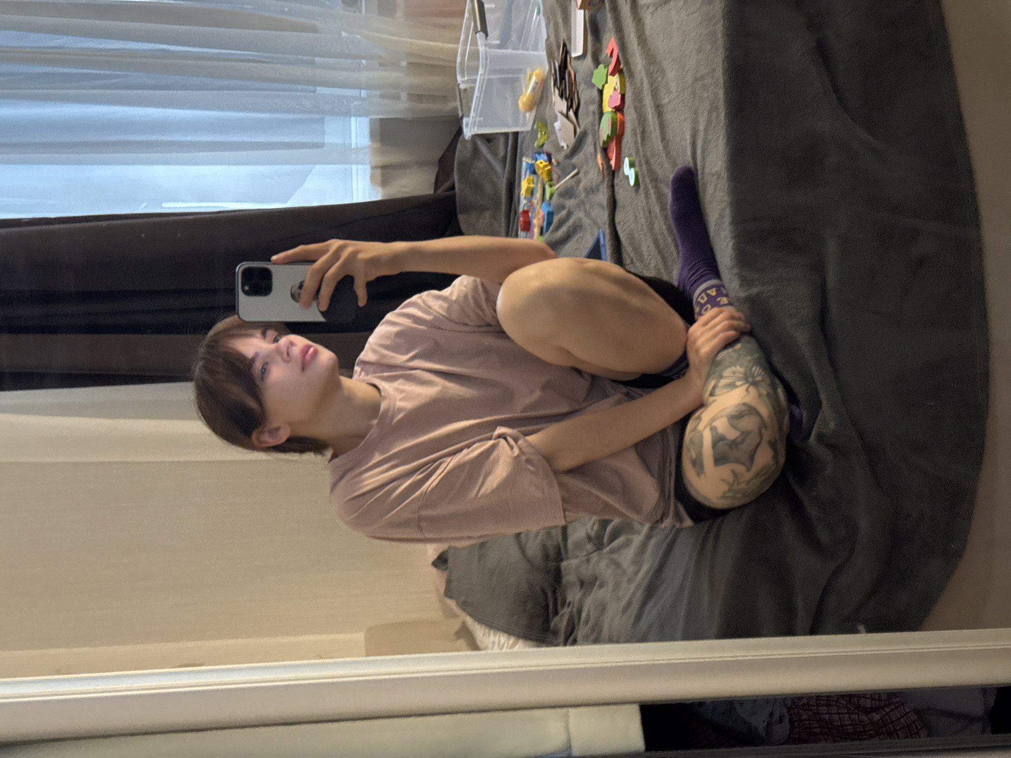 A person with short brown hair and light skin is sitting cross-legged on a gray bed, holding a smartphone in their left hand as if taking a selfie in a mirror. The person is wearing a pink t-shirt and a purple sock. Their right leg, resting on the bed, has a visible tattoo. Various small objects are scattered on the bed next to them, including what appears to be a clear plastic container and colorful magnetic letters or shapes. The background includes a window covered by sheer white curtains. The image is taken from an angle, suggesting it is a reflection in a mirror. There is no information to suggest a location in the city of Rostov-on-Don, Russia.
