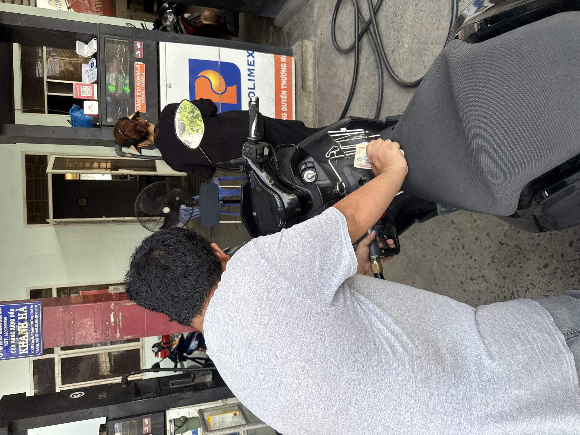 An over-the-shoulder shot captures a man in a light gray t-shirt and jeans at a gas station, holding a fuel nozzle and a stack of Vietnamese Dong banknotes. He appears to be either paying for fuel or preparing to do so, with the banknotes in his hand. His right hand is gripping the nozzle attached to a black motorcycle.
In the background, a woman wearing a black top and a visor stands at a gas pump, facing away from the camera. The gas pump displays "XĂNG E10 RON95-III". To the left of the man, a blue sign with Vietnamese text reads "CÔNG TY TNHH VẬN TẢI - XĂNG DẦU CƯỜNG THỊNH MST: 4200356850" and below that "KHÁNH HÀ". The location indicated on the sign is "DC: 52/54 Hùng Việt, P. Tân Lập, TP. Nha Trang, T. Khánh Hòa". This confirms the location is a gas station in Bac Nha Trang Ward, Vietnam.
Other elements in the scene include a black motorcycle, a fan, and various signs and equipment associated with a gas station. The ground is paved with concrete and gravel.